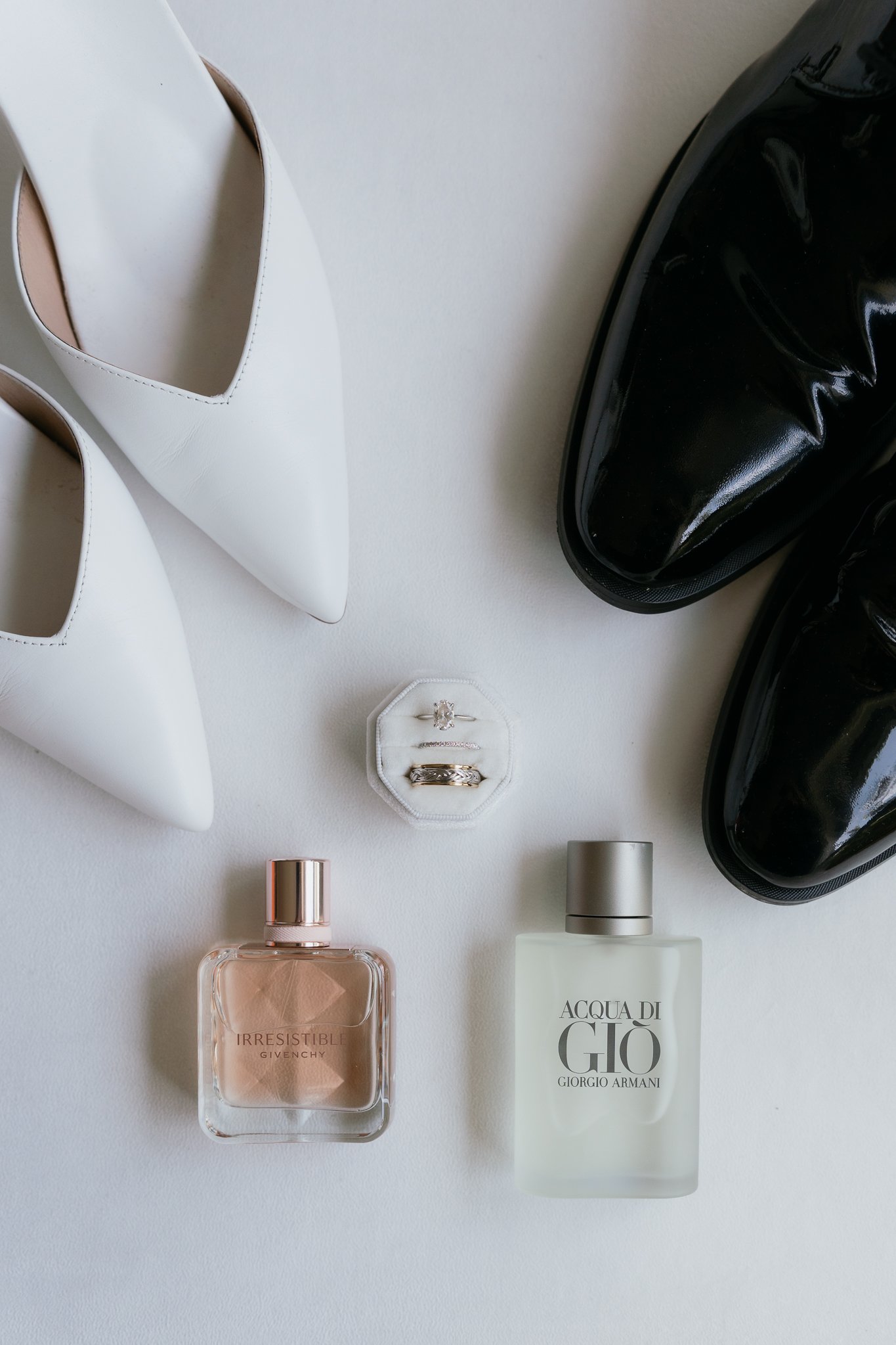 A flat lay of fashion and beauty items on a white surface, including white high-heeled shoes, black shiny boots, a white ring box with rings, two perfume bottles labeled 'Irresistible' by Givenchy and 'Acqua di Giò' by Giorgio Armani, and a small lipstick and lotion container.