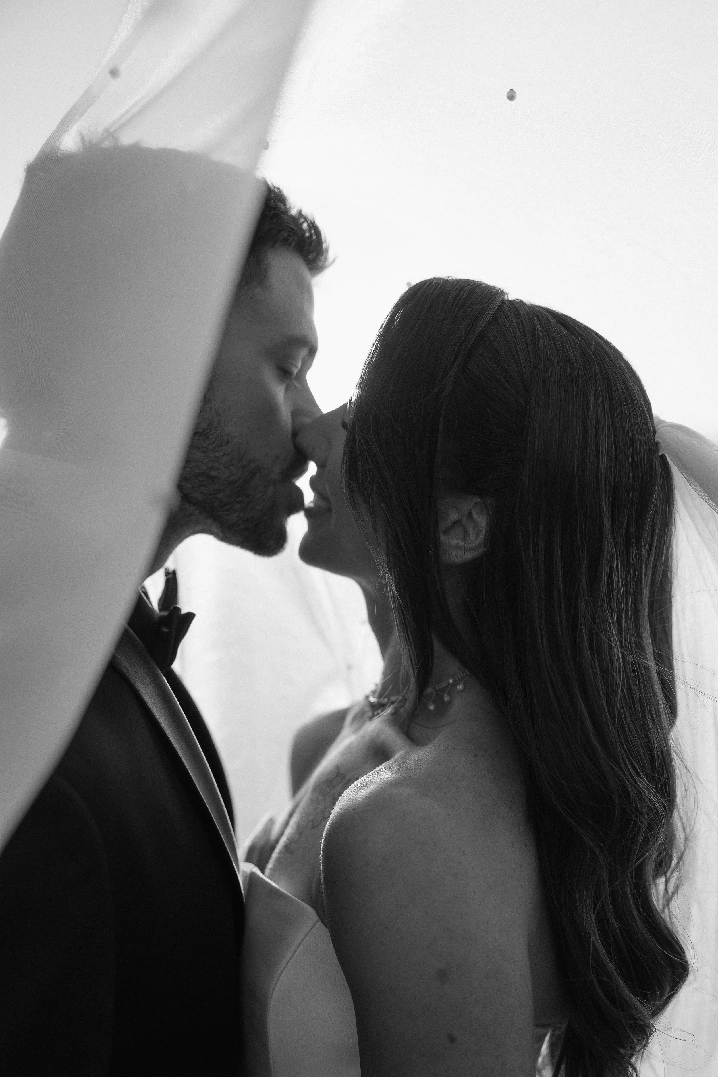 Black and white photo of a couple kissing with their noses touching; the man has short hair and a beard, wearing a tuxedo, and the woman has long hair, a necklace, and is in a strapless dress.