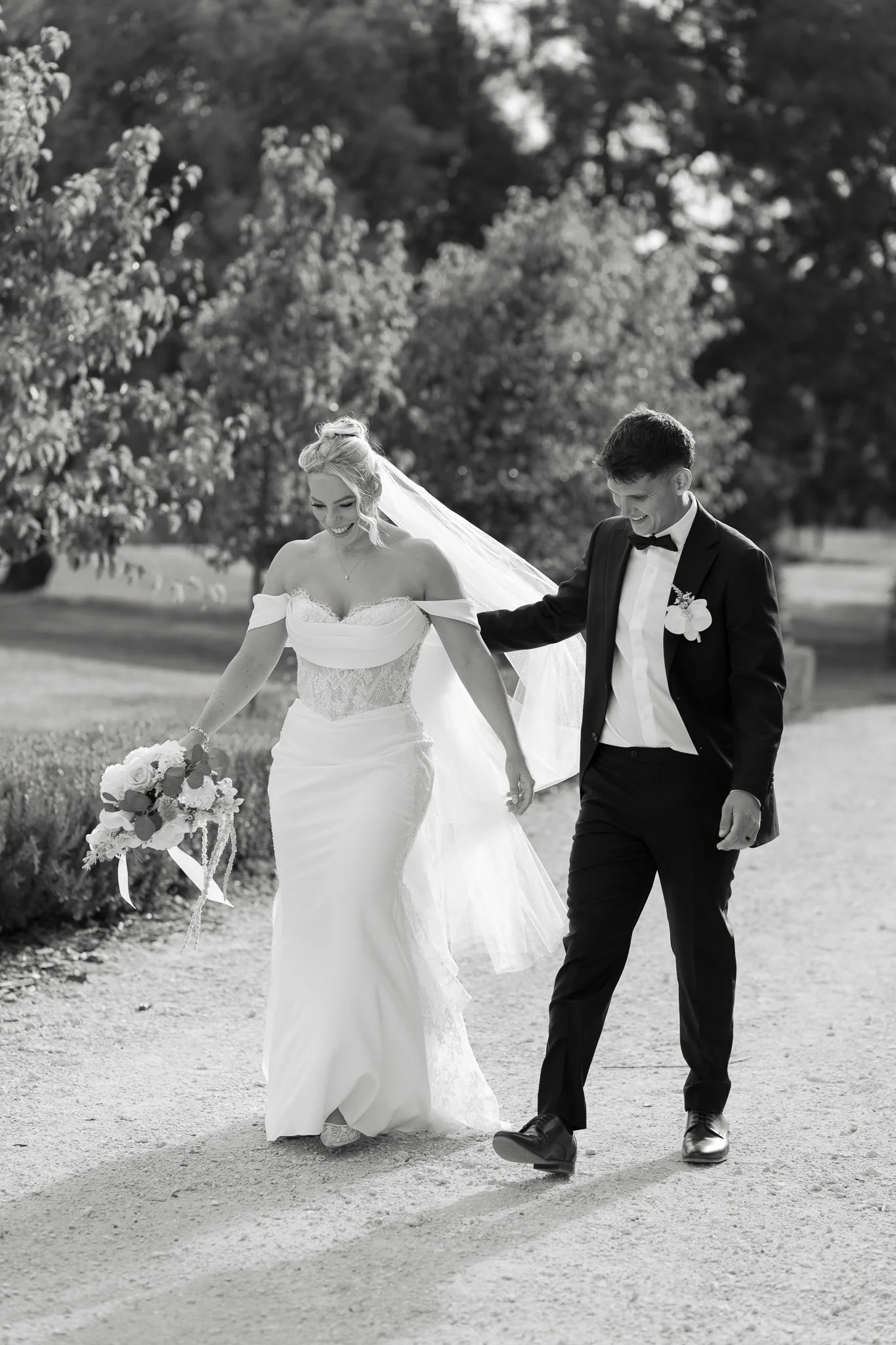 A black and white photo of a bride and groom walking hand in hand outdoors, smiling and looking happy. The bride is wearing a lace wedding gown and holding a bouquet, while the groom is in a tuxedo.