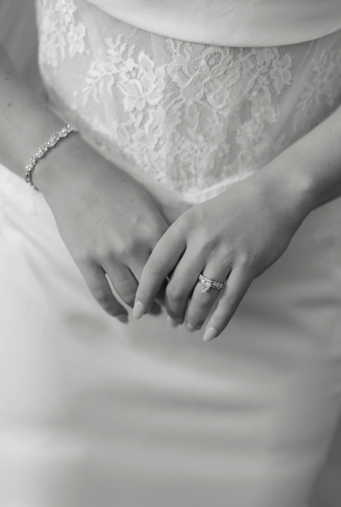 Close-up of a woman's hands showing an engagement ring and bracelet, with a lace dress in the background in black and white.