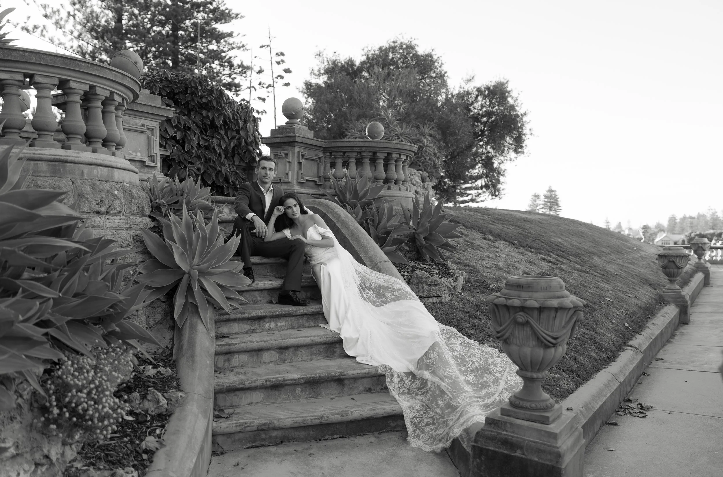 A man in a suit and a woman in a wedding dress sitting on stone steps in a garden, surrounded by plants and historic stone architecture, with a scenic view of trees and open land in the background.