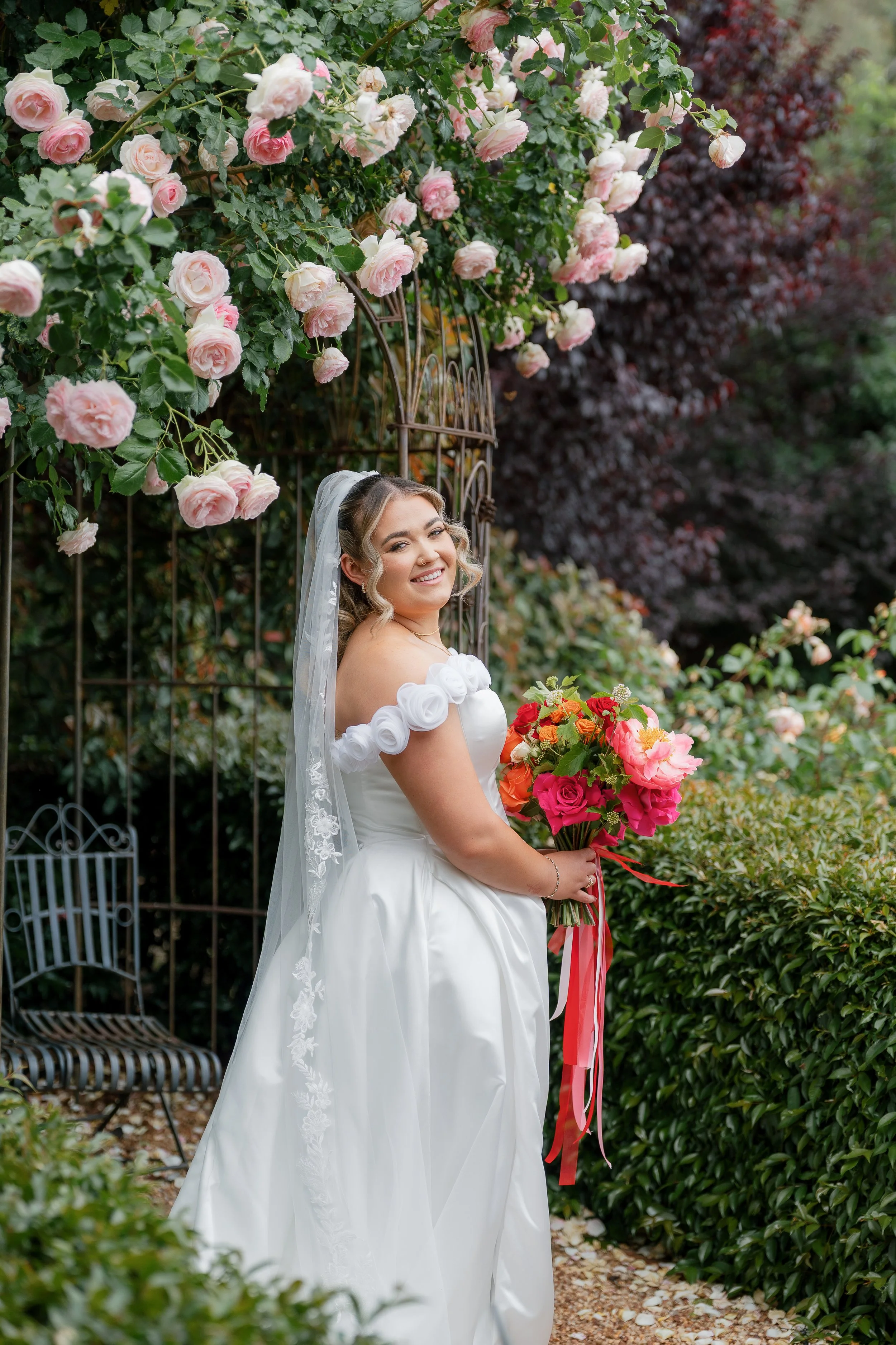 A bride in a white wedding gown holding a bouquet of pink and orange flowers, standing outdoors in front of a garden arch covered with pink roses, smiling and looking at the camera.