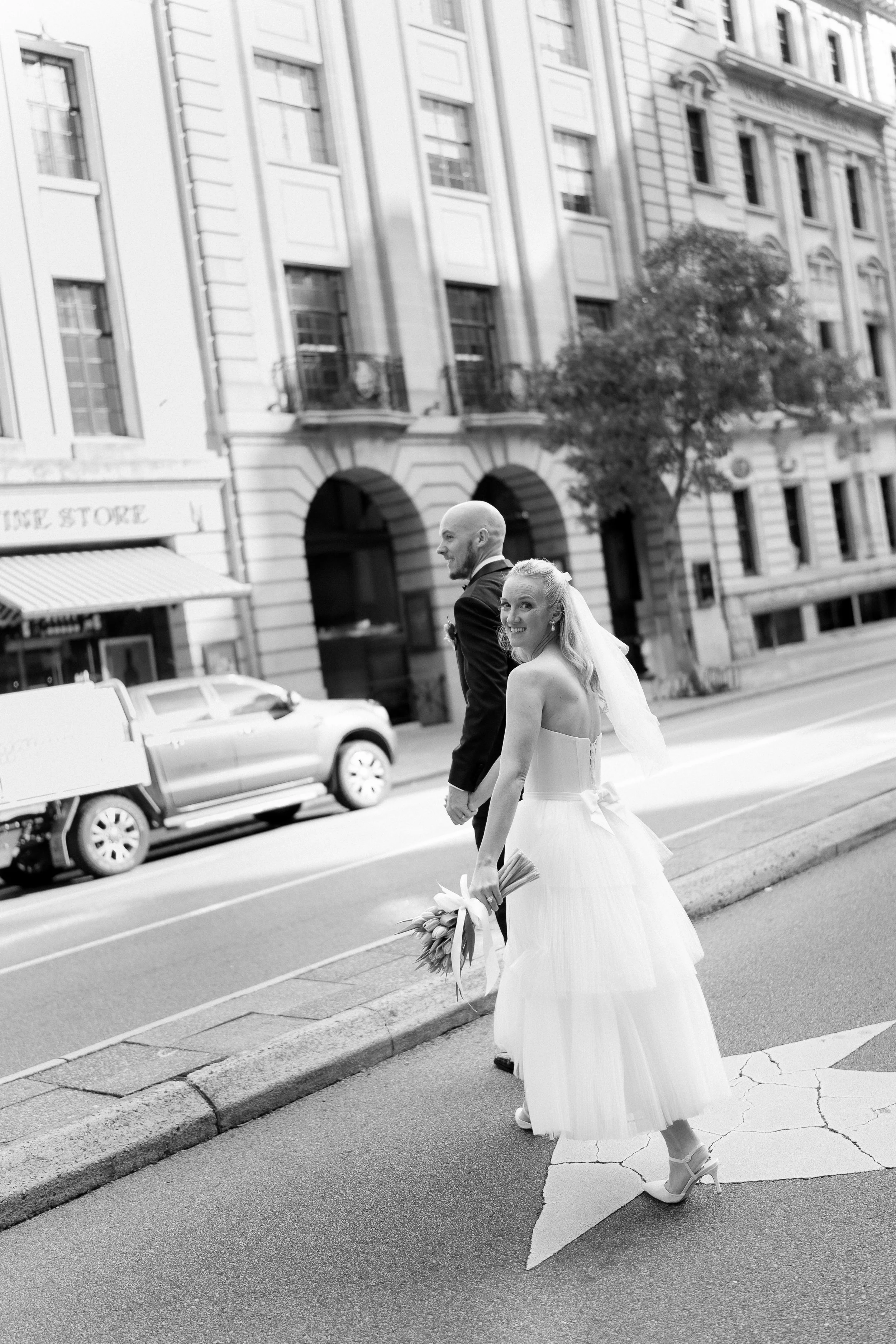 A bride and groom walking on a city street, with the bride holding a bouquet, smiling at the camera, and the groom walking beside her, both dressed in wedding attire.