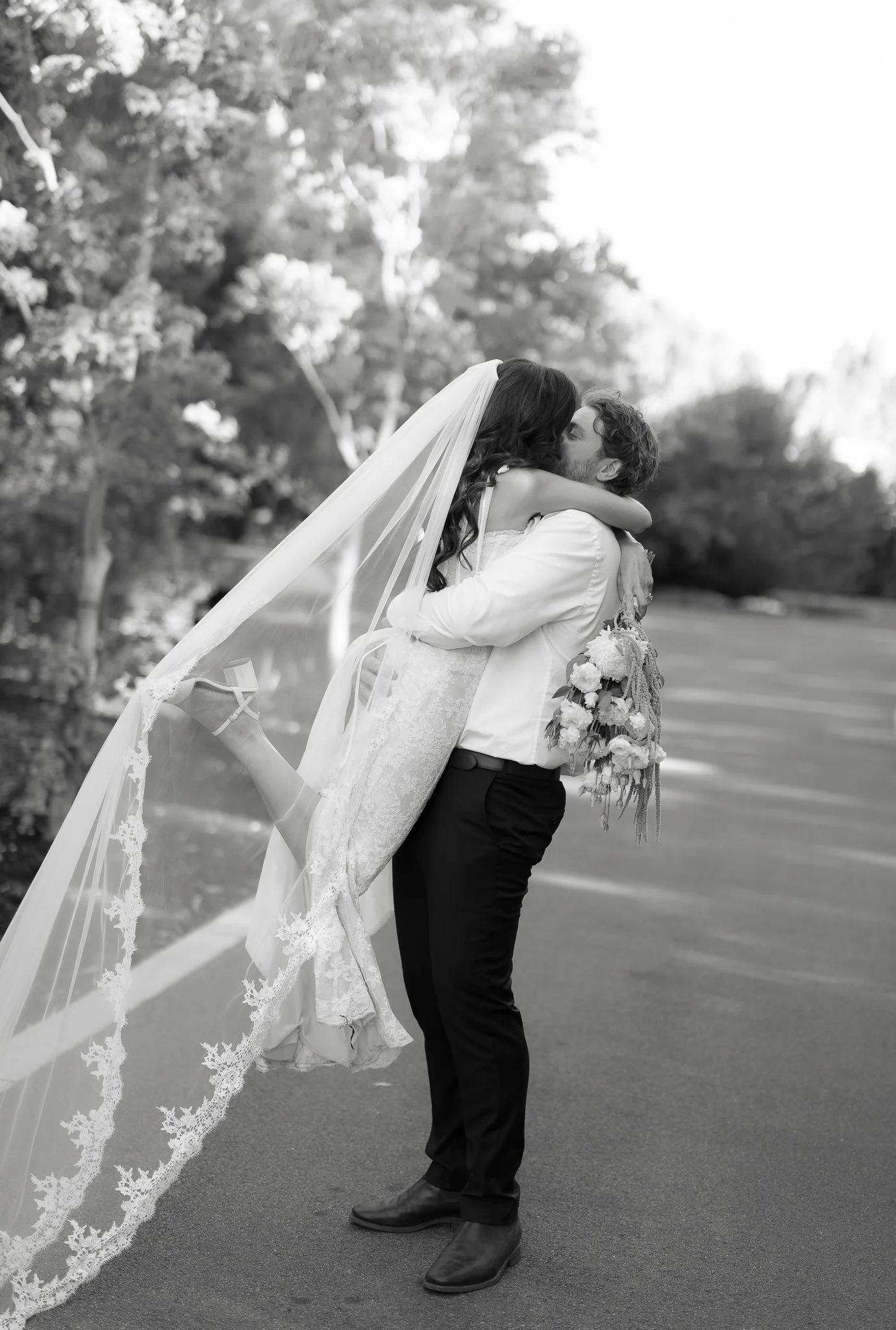A couple sharing a kiss outdoors, with the woman in a wedding dress and veil, and the man in a white shirt, black pants, and black shoes. The woman is hugging the man, who is holding a bouquet of flowers.