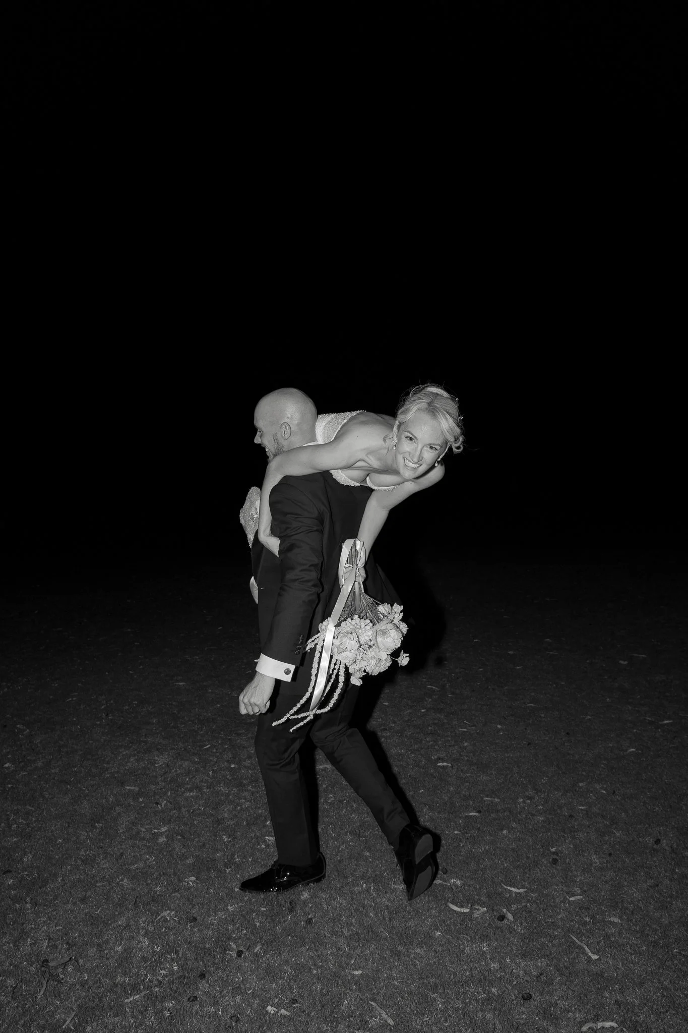 Black and white photo of a smiling woman with curly hair being carried on the back of a man in a tuxedo, holding a bouquet of flowers, against a dark background.