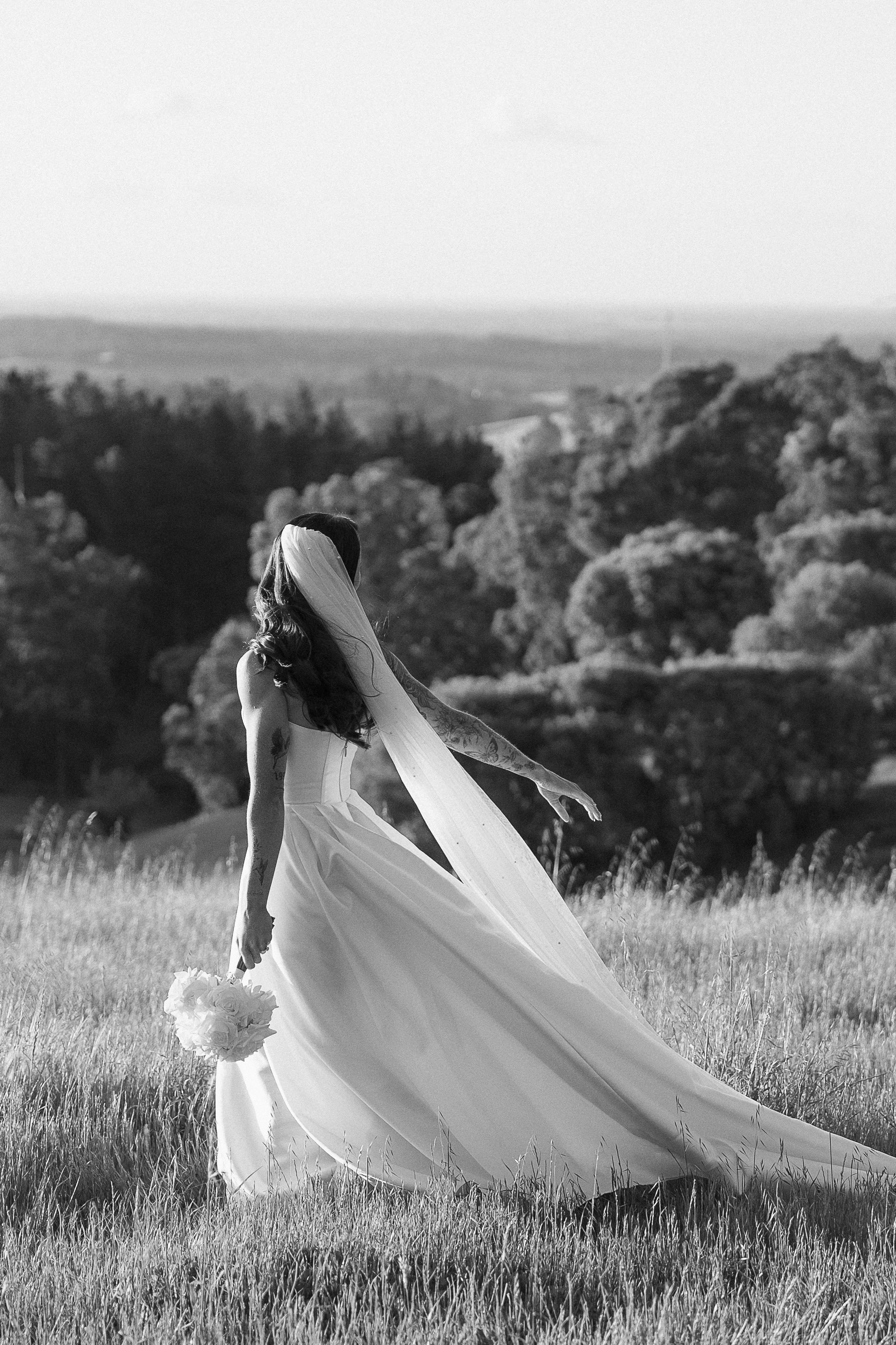 A bride standing in a grassy field wearing a wedding gown, holding a bouquet, with tattooed arms, and a veil flowing in the wind, overlooking a landscape with trees.