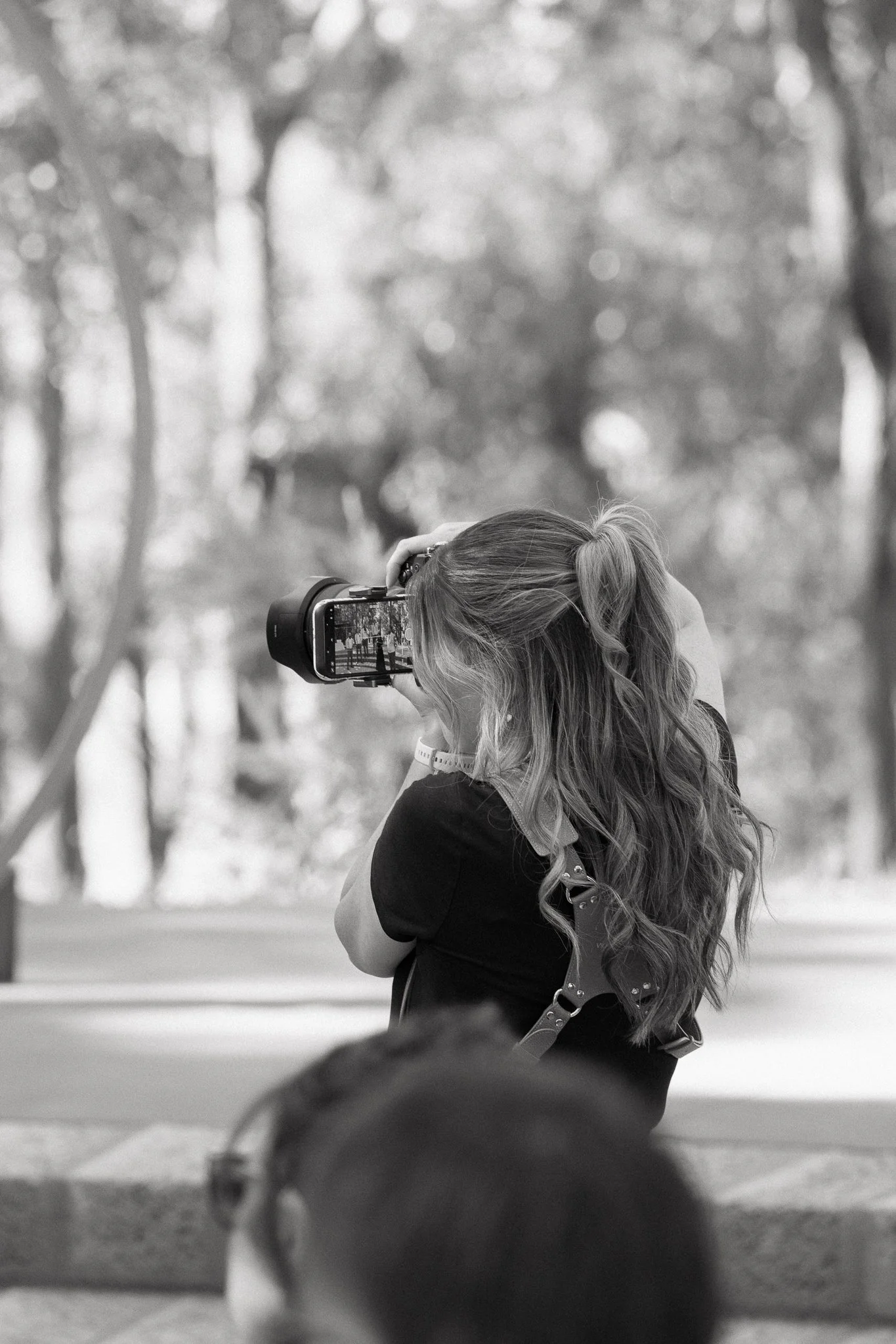 A young woman with long, wavy hair, wearing a black shirt and a watch, taking a photo with a camera in an outdoor setting with trees in the background.