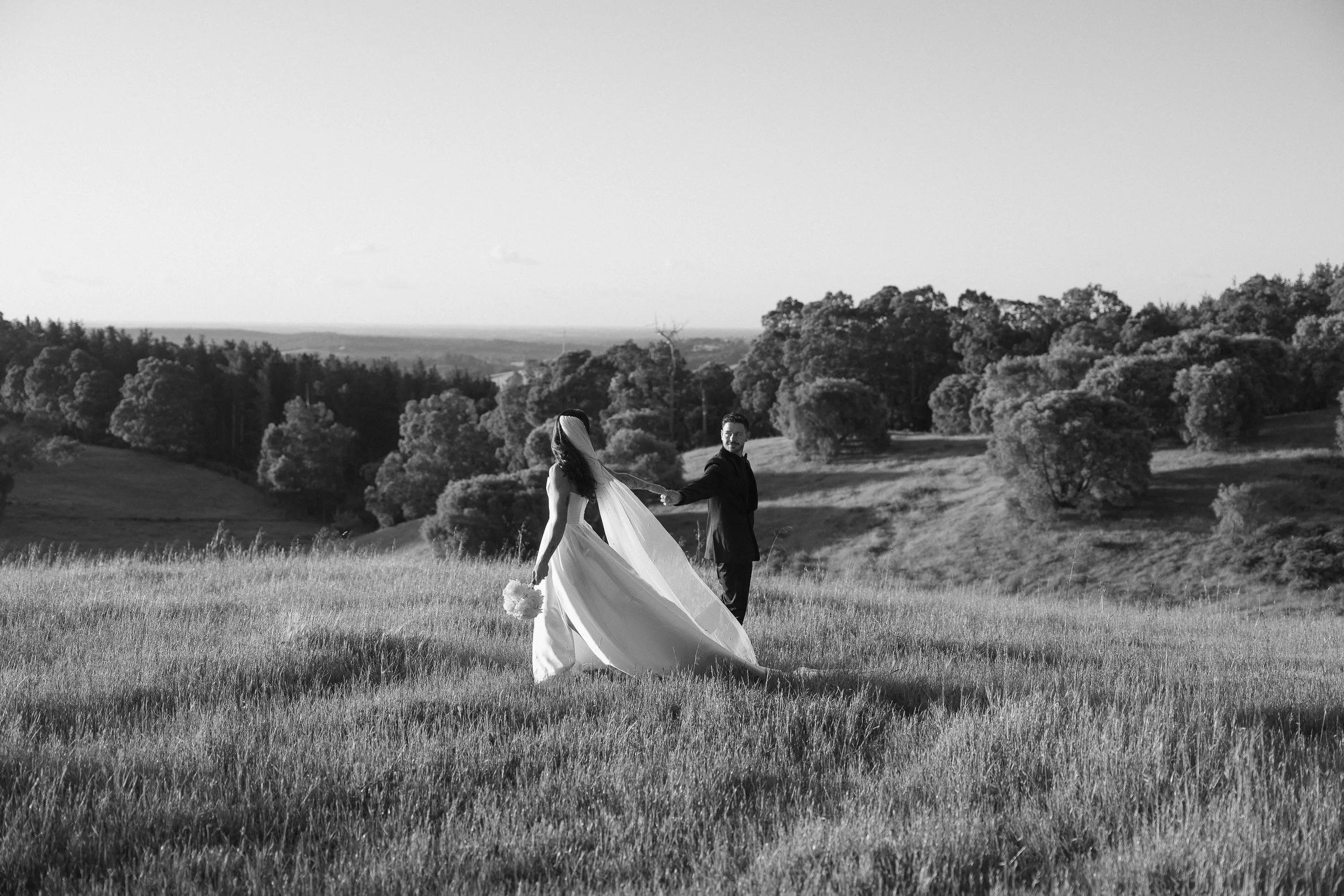 A bride and groom holding hands in a field with trees and hills in the background, black and white photograph.