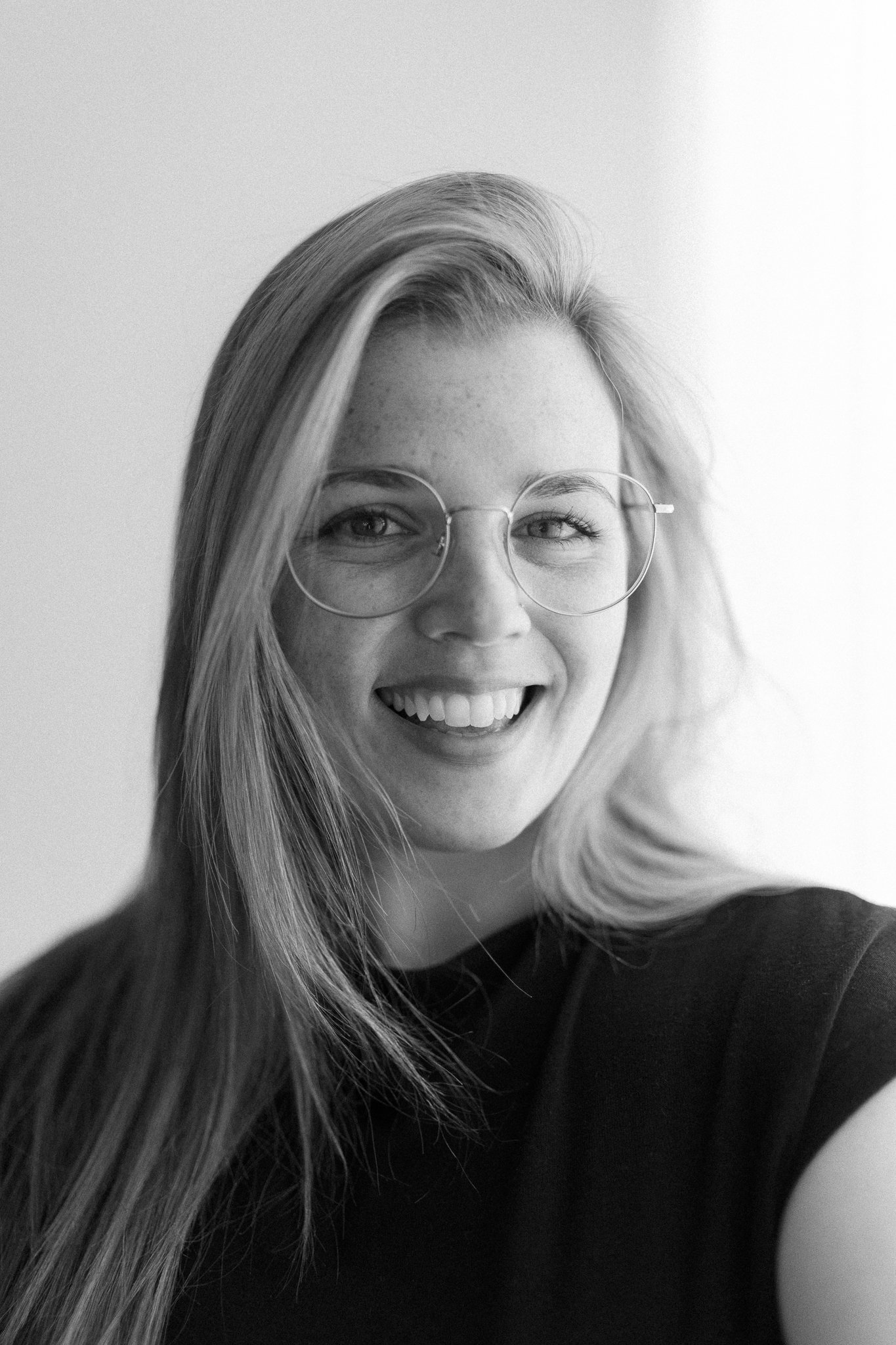 A smiling young woman with glasses and long hair, wearing a black shirt, in black and white photo.