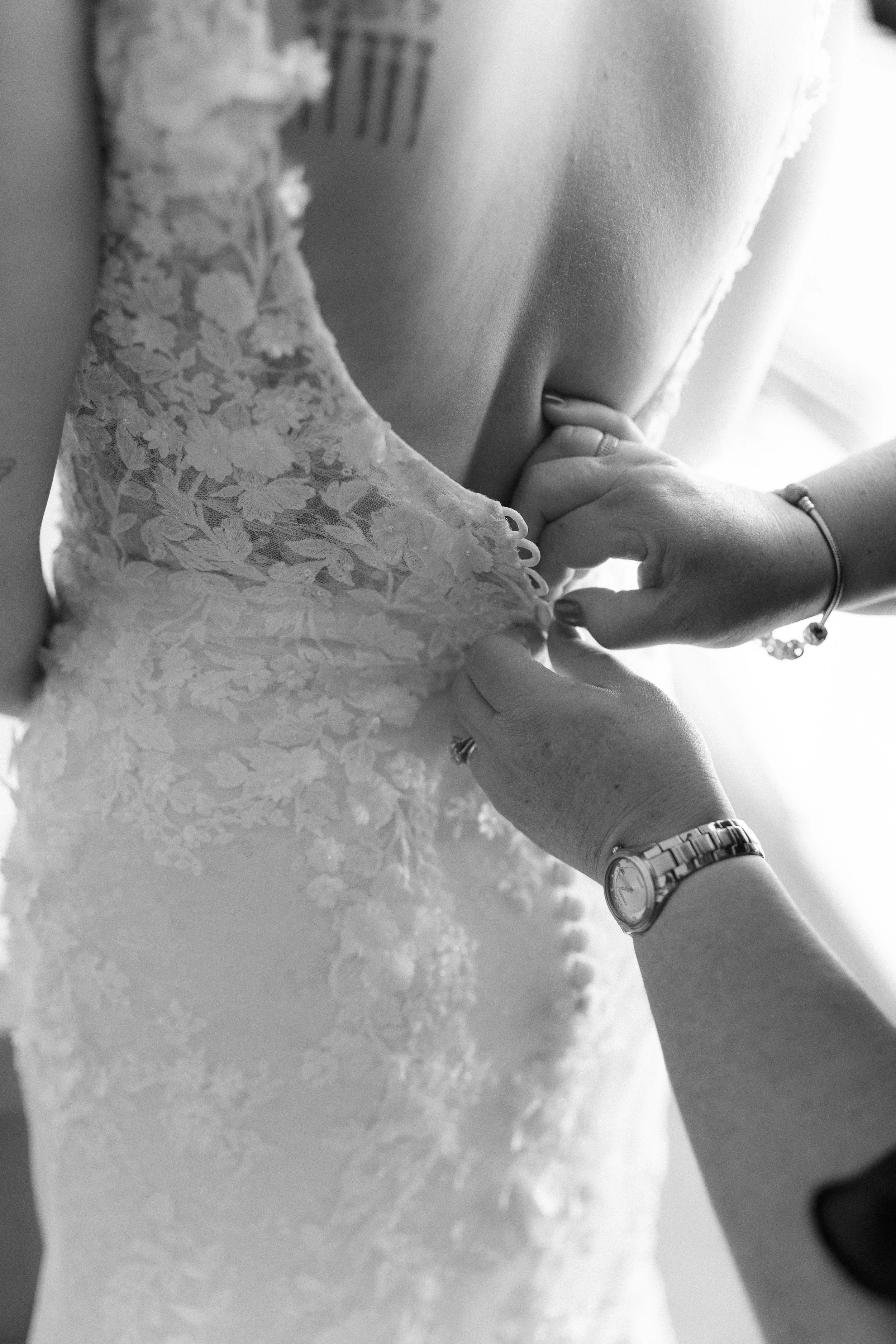 A person is buttoning the back of a wedding dress with lace details, while wearing a watch and rings.