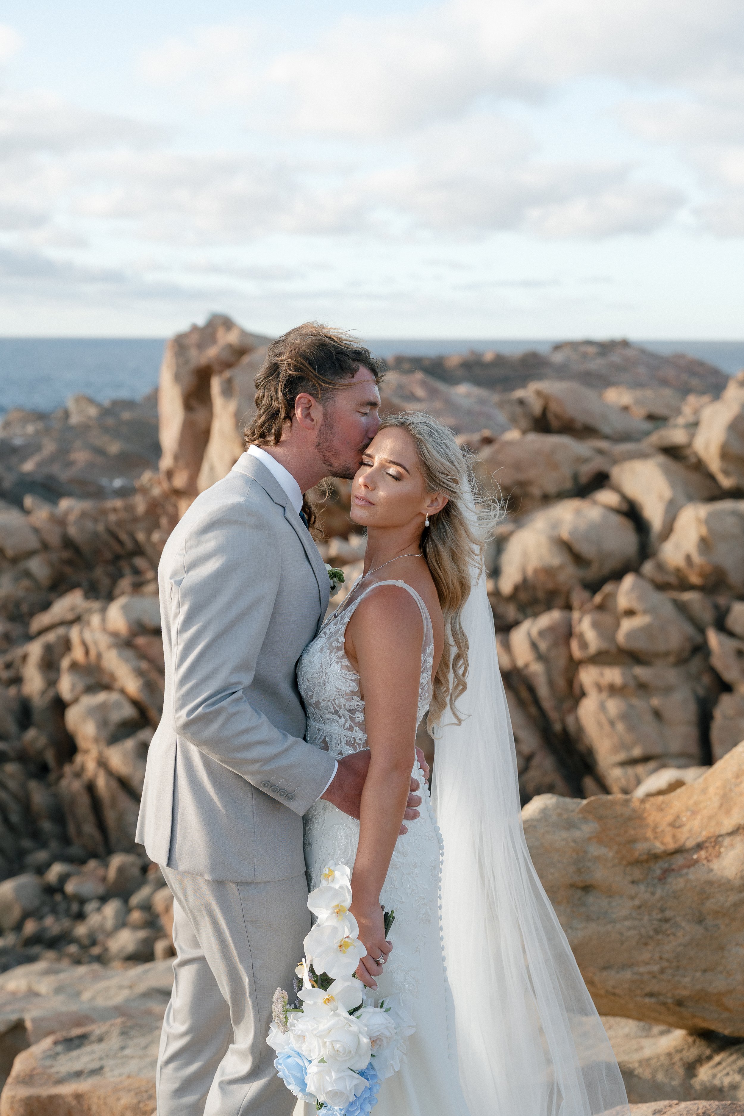 A newlywed couple stands close on a rocky seaside, the groom gently kisses the bride's forehead, she holds a bouquet of white flowers, both in elegant wedding attire with ocean in the background.
