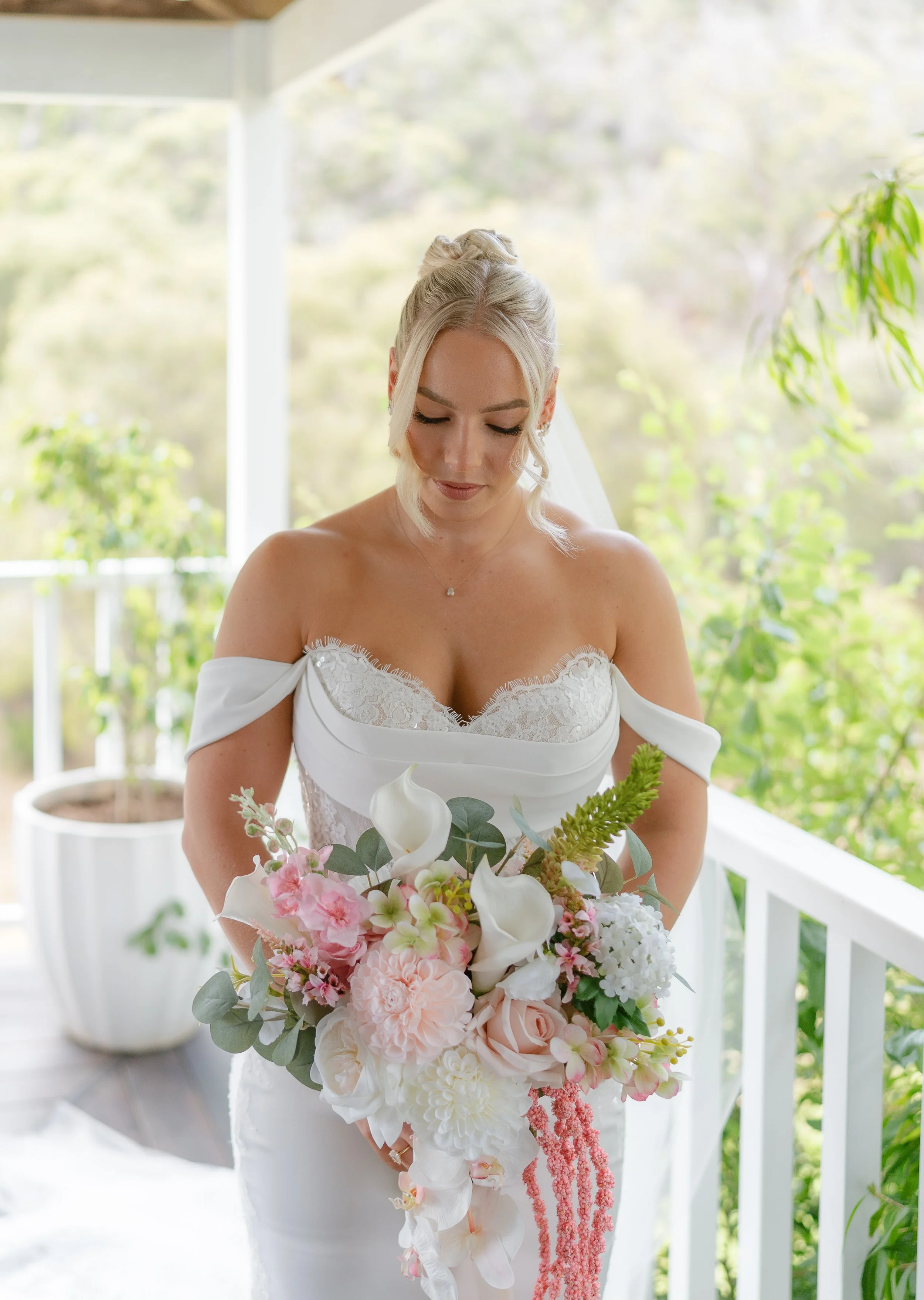 A woman in a white wedding dress holding a large bouquet of pink and white flowers on a porch with greenery in the background.