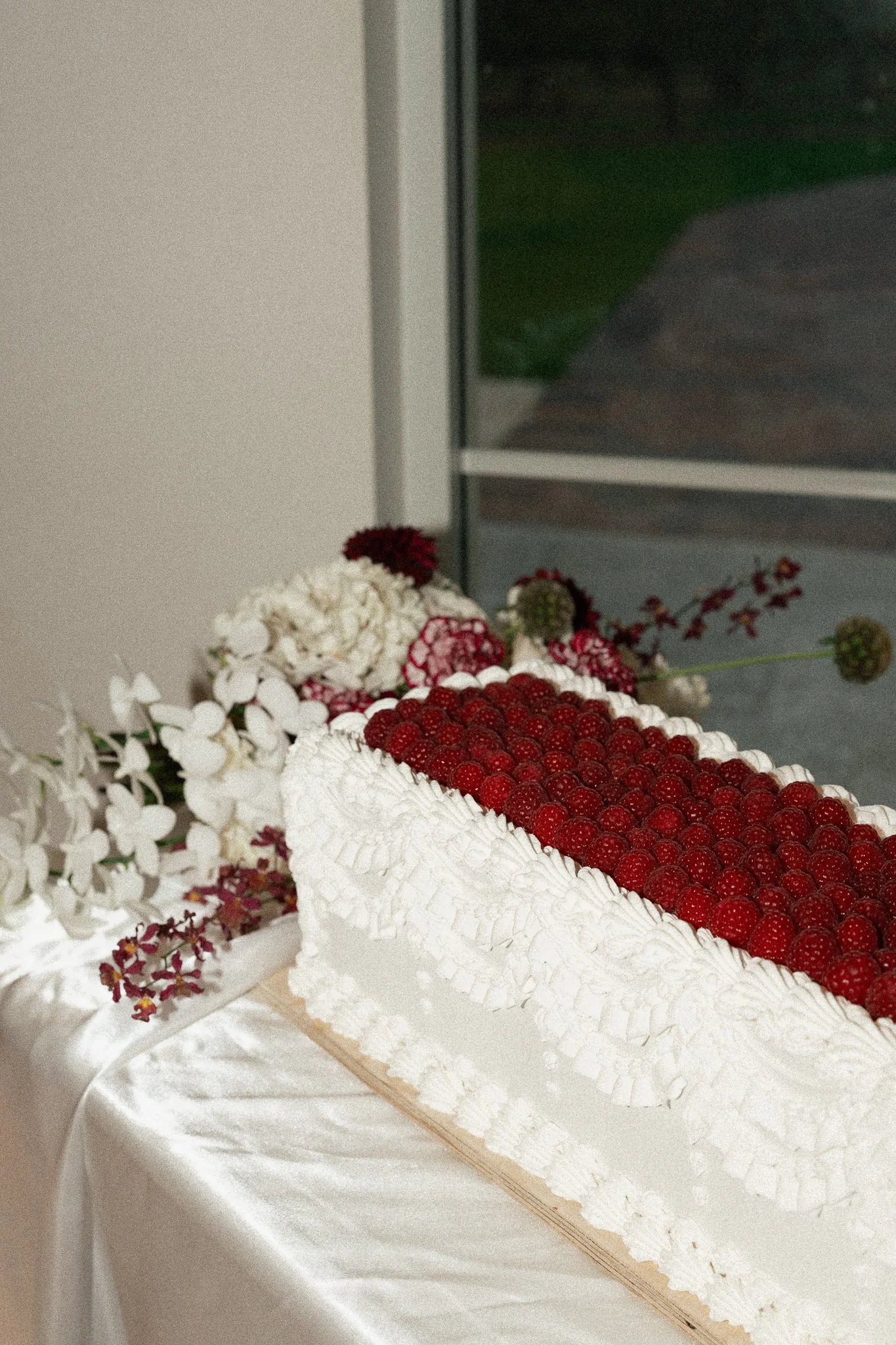 A wedding cake decorated with fresh red raspberries on top and white icing with floral accents on the sides, placed on a white tablecloth.