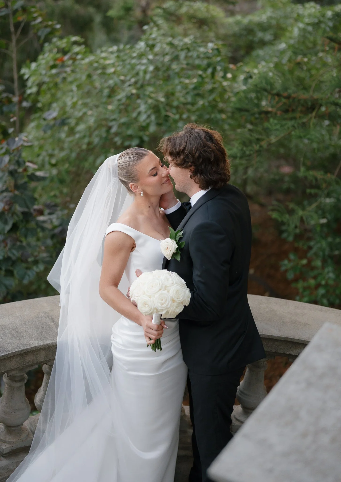 A bride and groom share a romantic moment, standing outdoors with green foliage in the background. The bride is wearing a white wedding gown and veil, holding a bouquet of white roses. The groom is dressed in a black tuxedo, gently touching the bride's face.