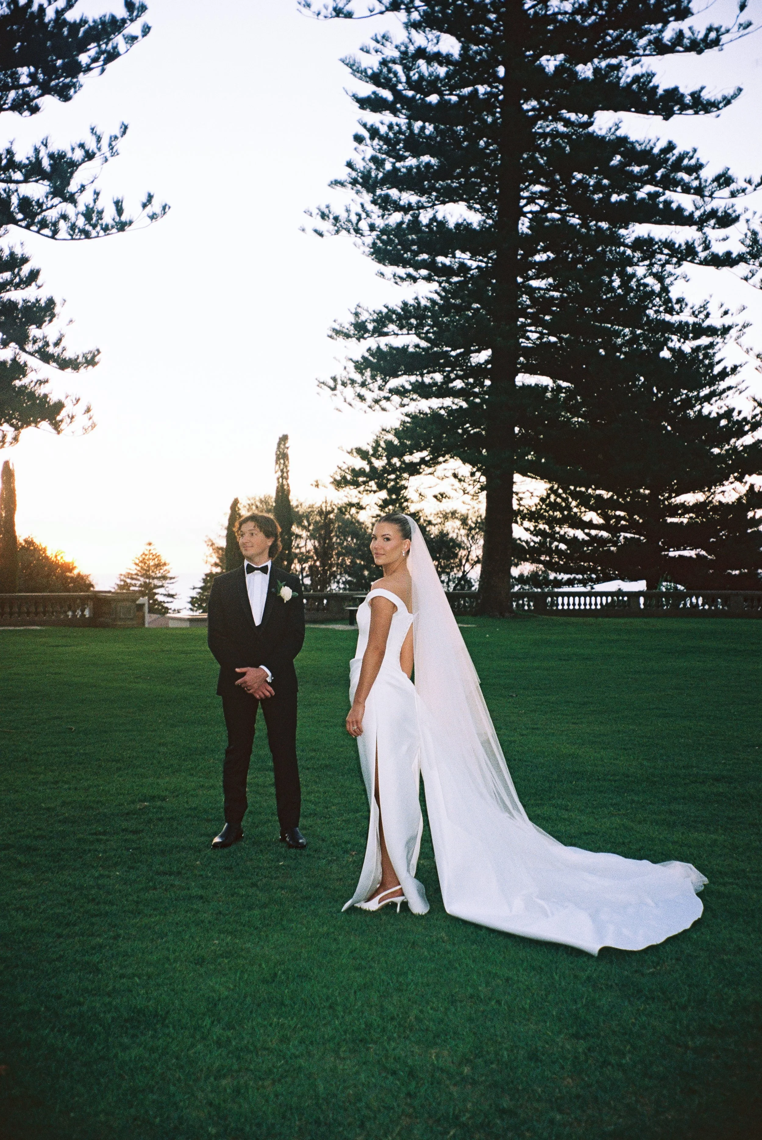 A bride and groom standing on a grassy area outdoors during sunset, with tall pine trees and a stone balustrade in the background. The groom is wearing a black tuxedo with a white shirt and a black bow tie, while the bride is wearing a white wedding gown with a long train, open back, veil, and high heels.