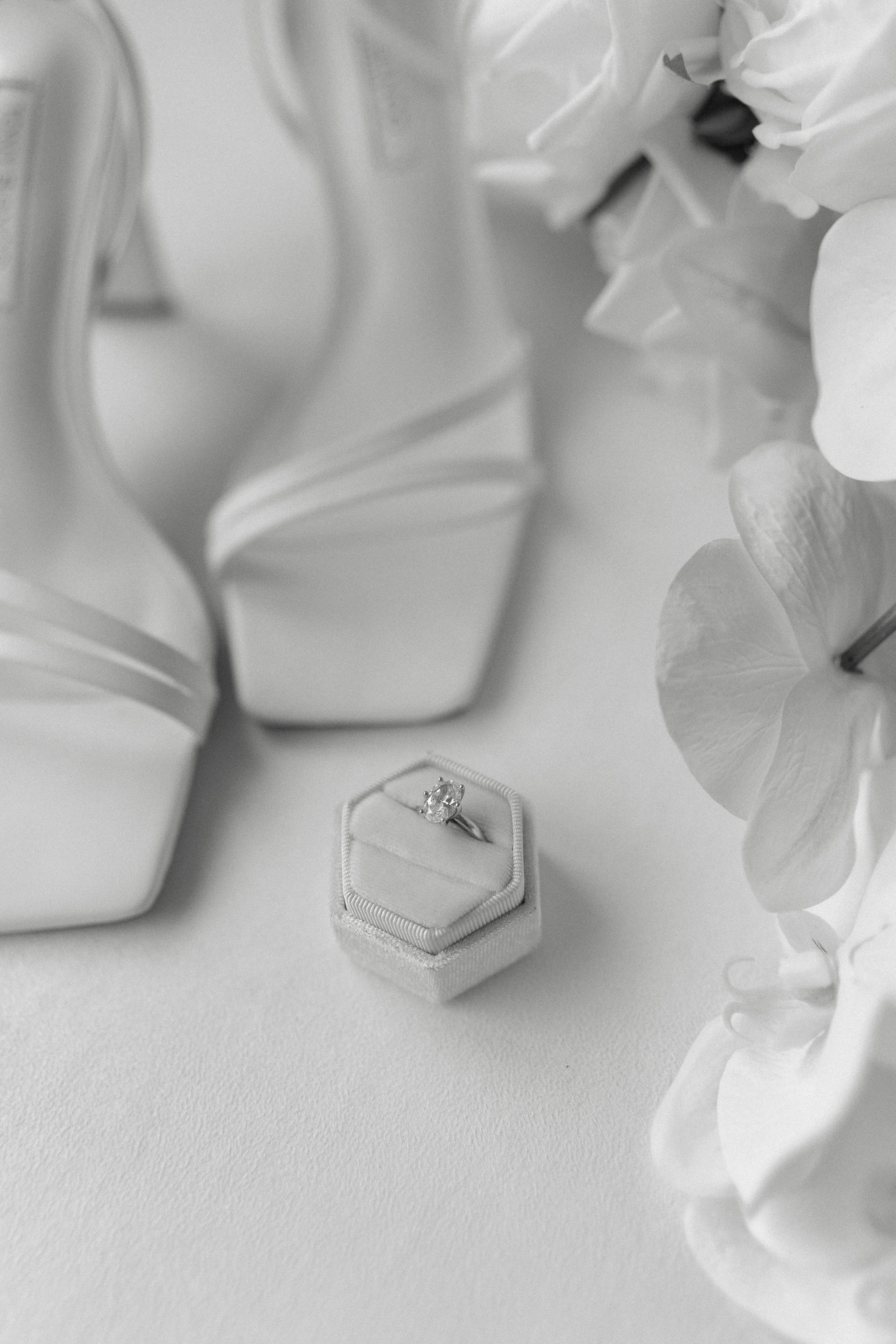 A diamond engagement ring in a small hexagonal box, with vases and flowers in the background, in black and white.