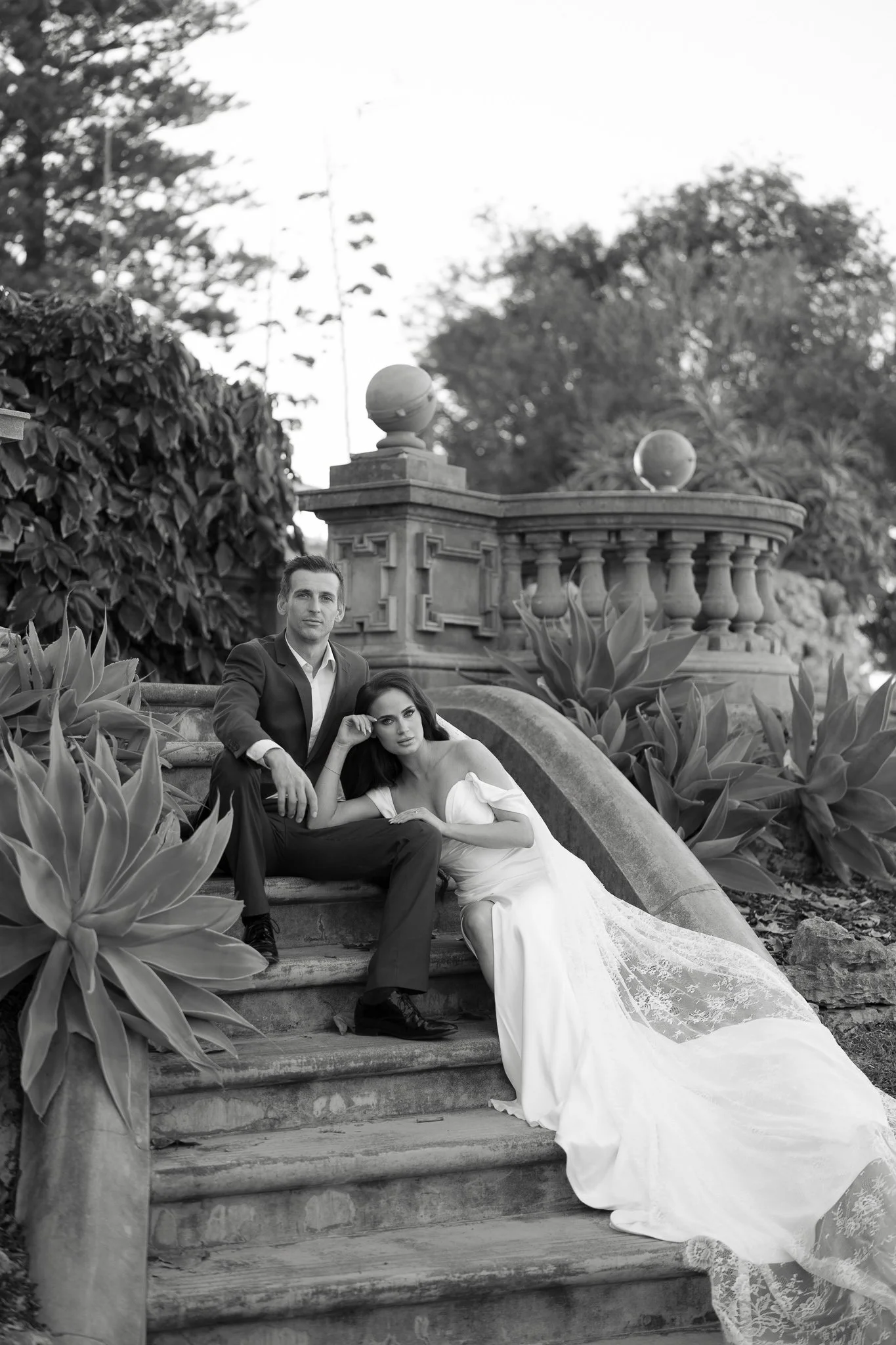 A man in a suit and a woman in a wedding dress sitting on stone stairs in a garden with lush plants and trees, posing for a photo.