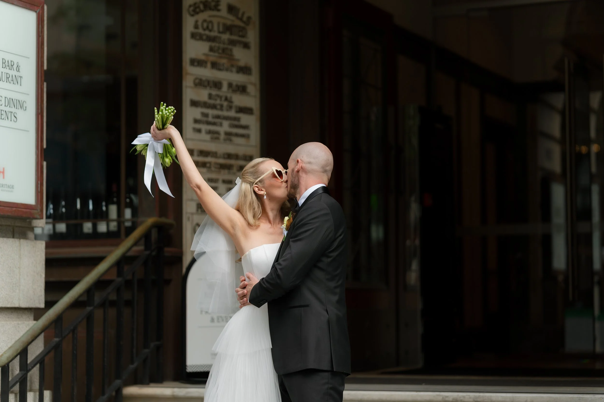 A bride and groom kissing outdoors, with the bride holding a bouquet of flowers raised in the air.