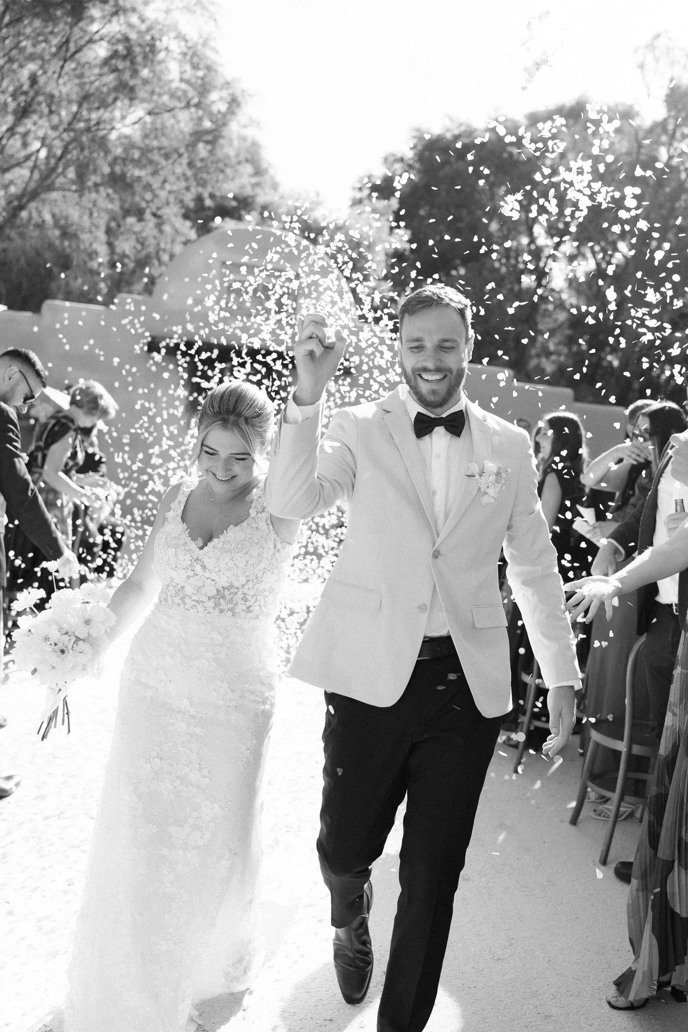 A bride and groom smiling and walking hand-in-hand during a wedding ceremony, with confetti falling around them.