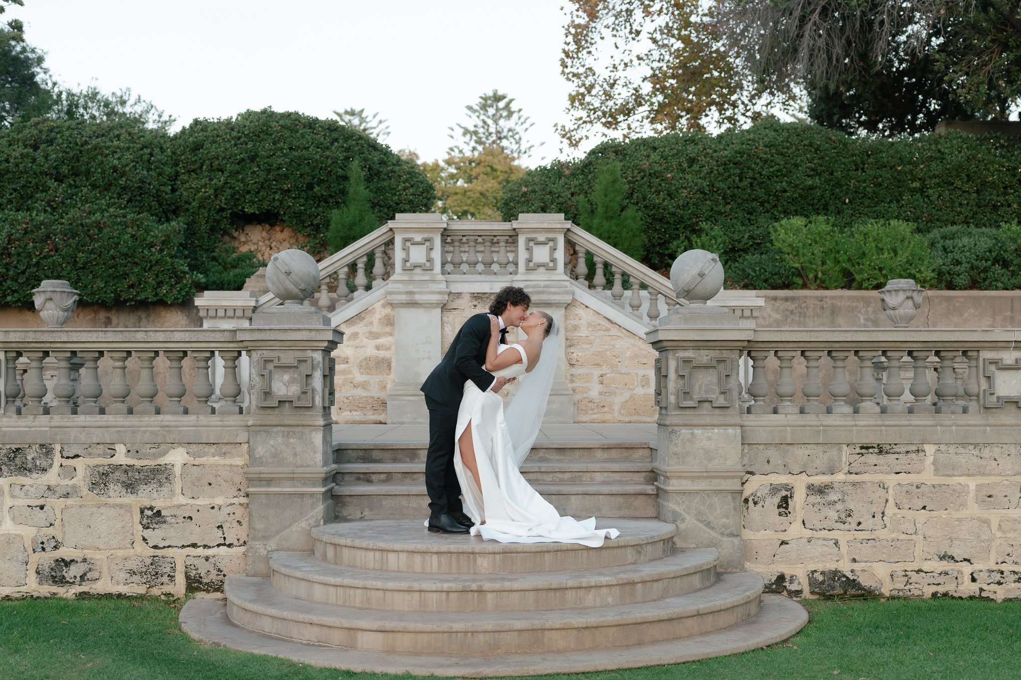 A bride and groom kissing on the steps of a stone and concrete outdoor staircase, with lush trees and bushes in the background.