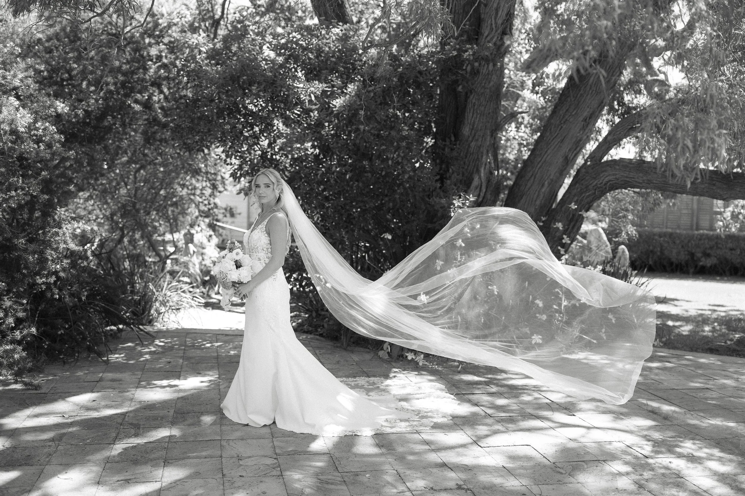 A bride in a wedding dress holding a bouquet of flowers, standing outdoors on a paved pathway under a large tree with a flowing veil trailing behind her.
