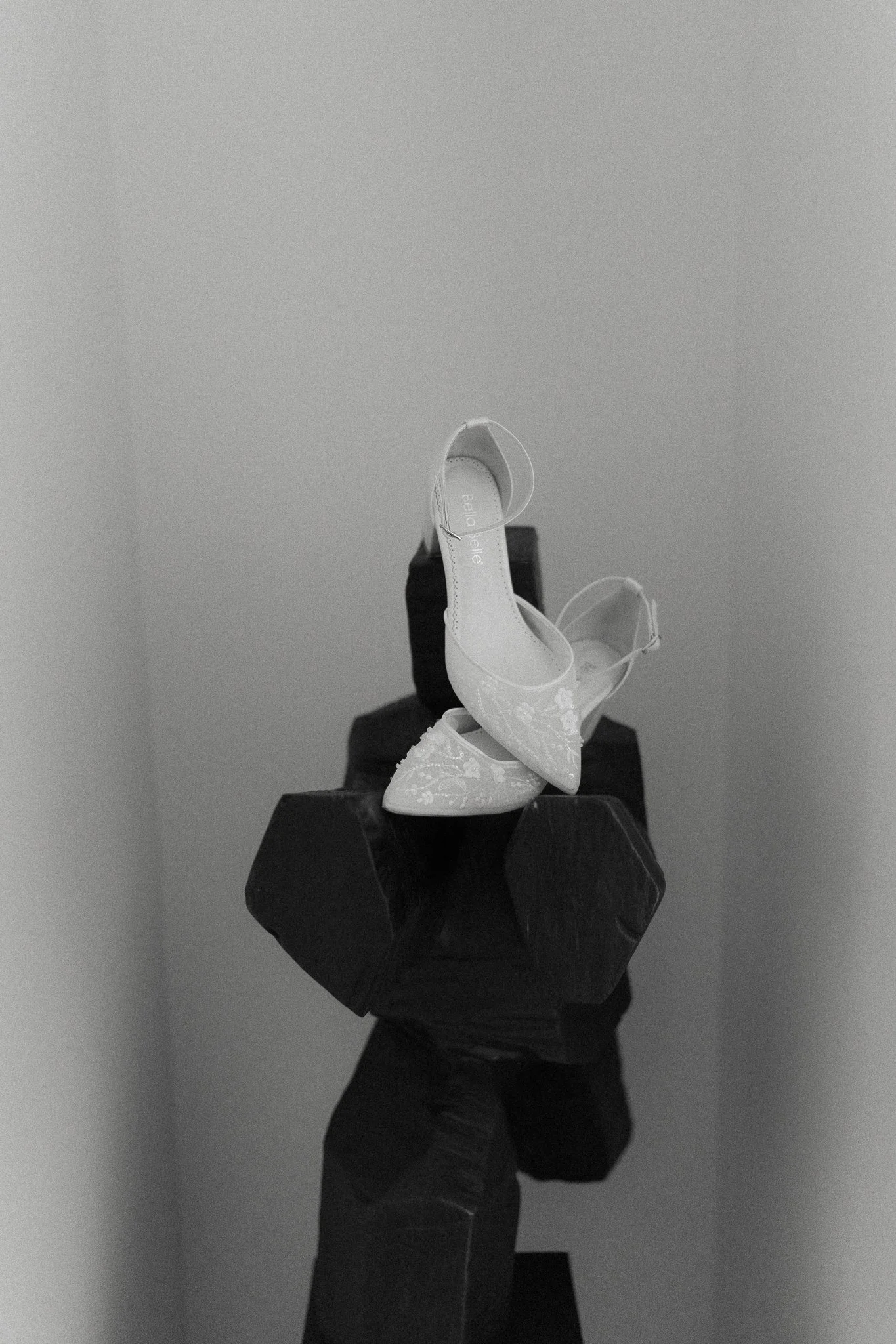 A pair of elegant high-heeled shoes with floral embroidery are balanced on a black, faceted sculpture or pedestal, all in black-and-white.