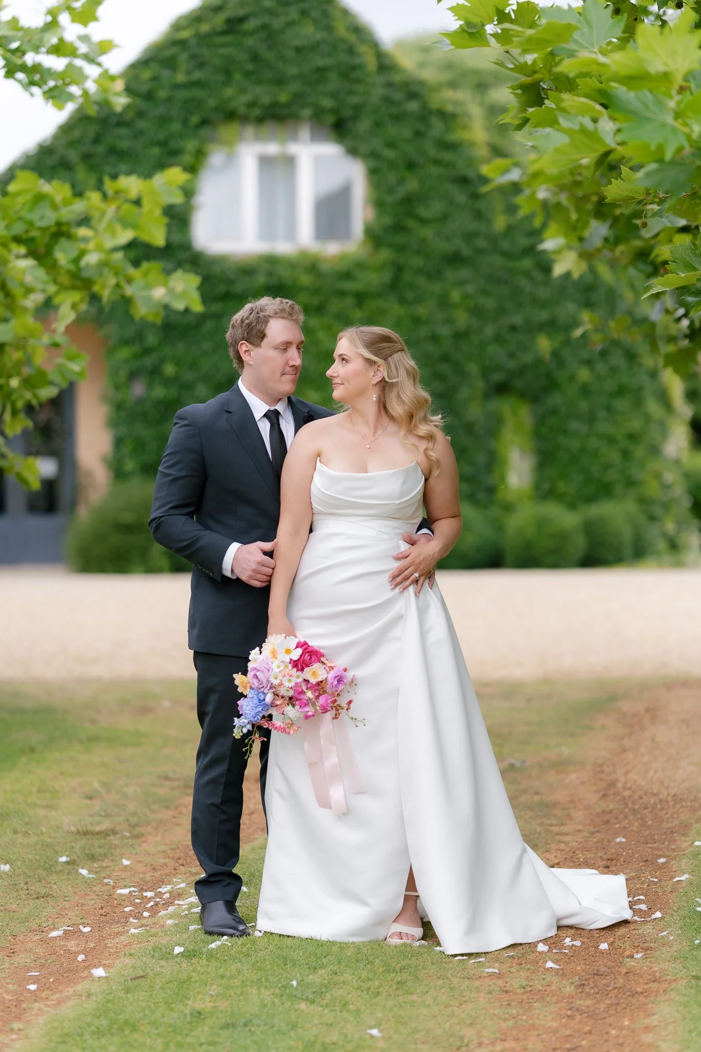 A bride and groom standing close together outdoors, with the bride holding a bouquet of pinks, purples, and whites, in front of a green ivy-covered building, surrounded by trees and falling flower petals.