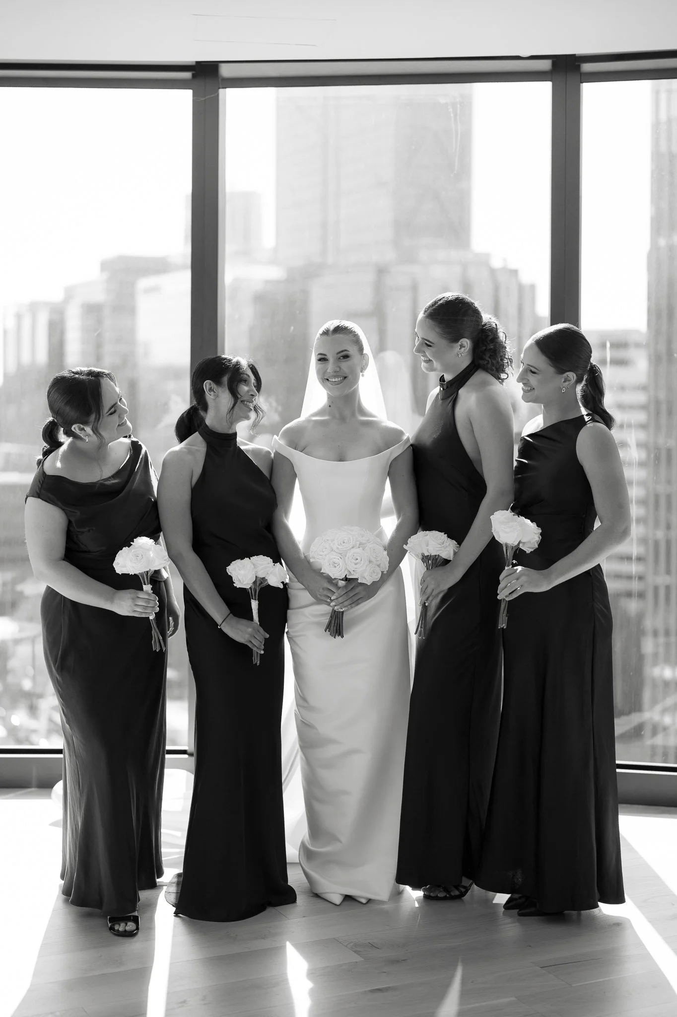 Black and white photo of a bride in a wedding dress standing in the middle with four bridesmaids in dark dresses holding bouquets, standing beside her in front of large windows with a city skyline in the background, smiling and looking at each other.