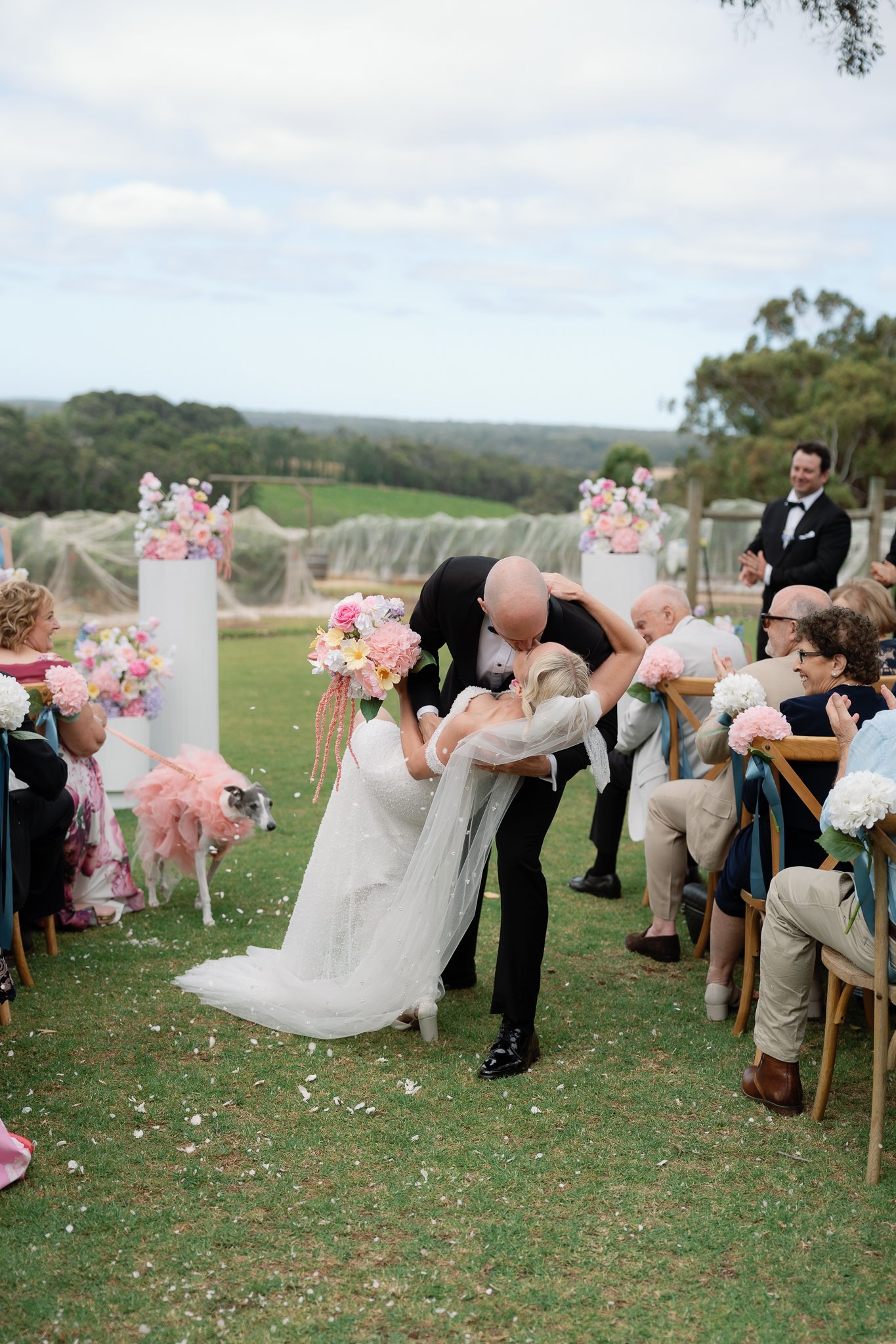 Bride and groom dancing at outdoor wedding, surrounded by seated guests with flowers, a dog in a pink tutu, and scenic countryside background.