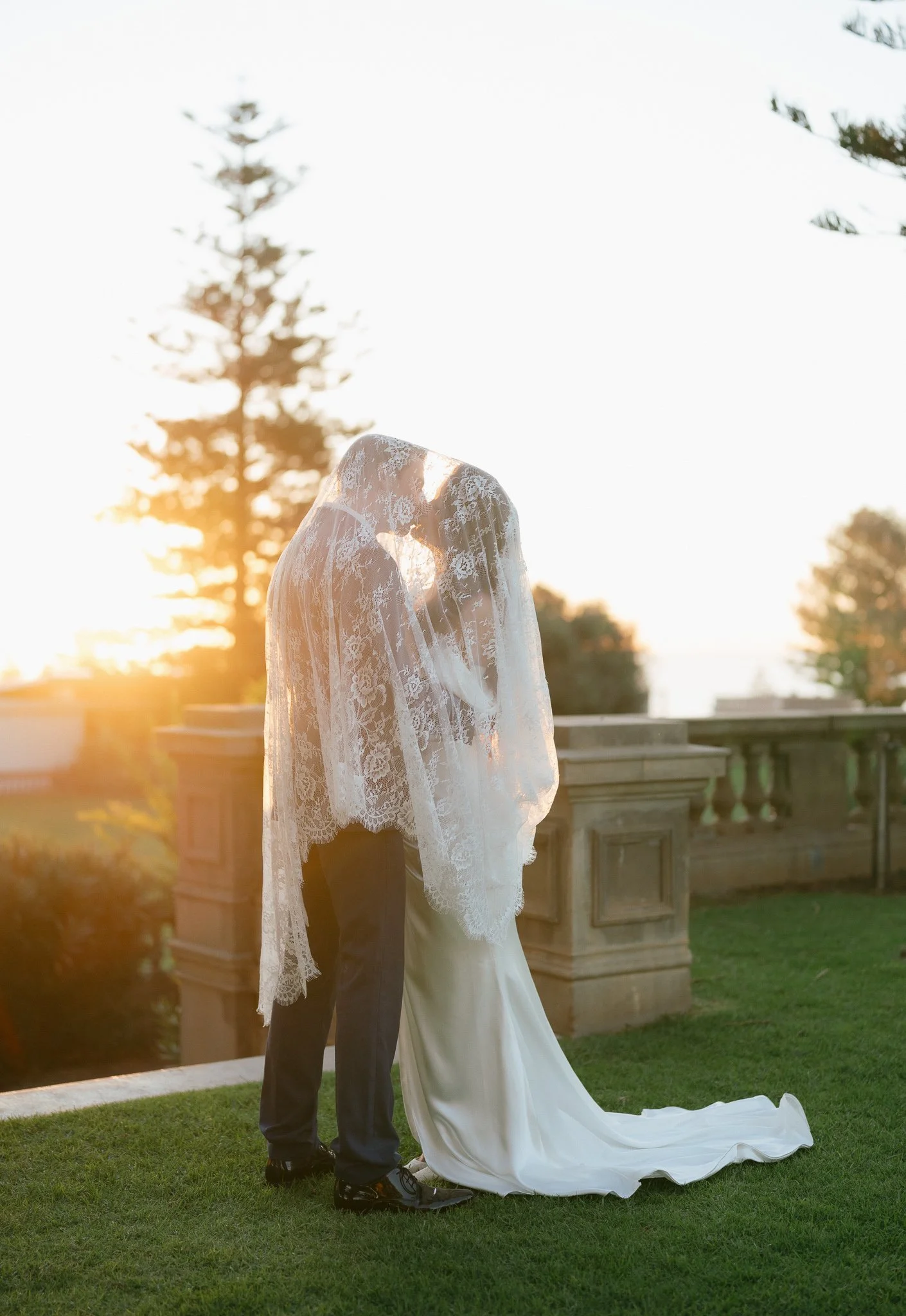 A bride and groom standing outdoors at sunset, embracing each other. The bride wears a white lace wedding dress with a long train, while the groom wears a dark suit. They are on a grassy area near a stone railing, with trees and the setting sun in th