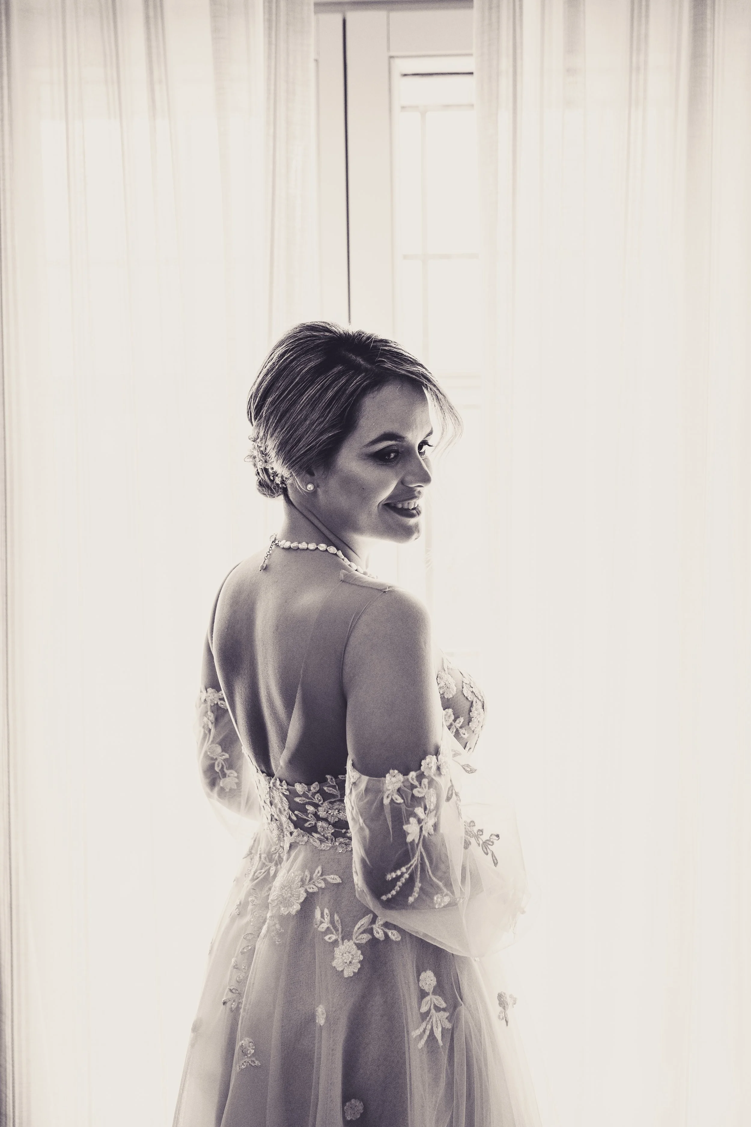 Mujer en vestido de novia con detalles de encaje, peinado recogido y collar de perlas, sonriendo frente a cortinas y ventana.