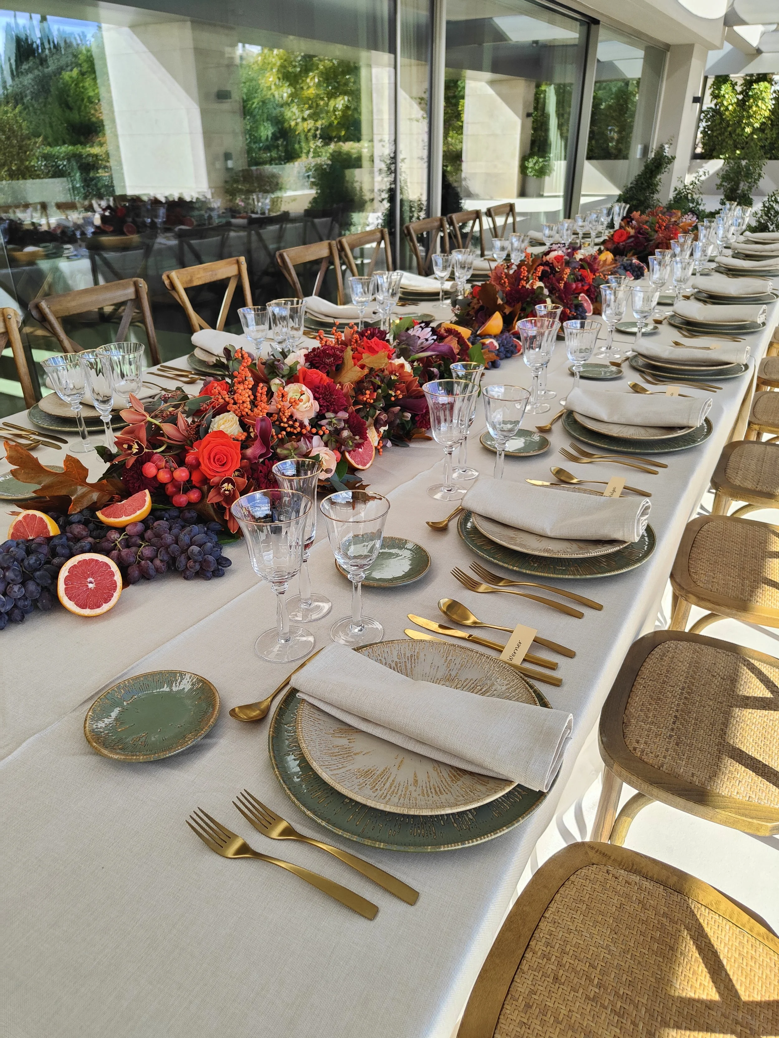 Mesa decorada para una celebración con un centro de flores en tonos cálidos, copas, platos, cubiertos de color dorado y servilletas blancas, en un espacio moderno con ventanas grandes y vistas a un jardín.