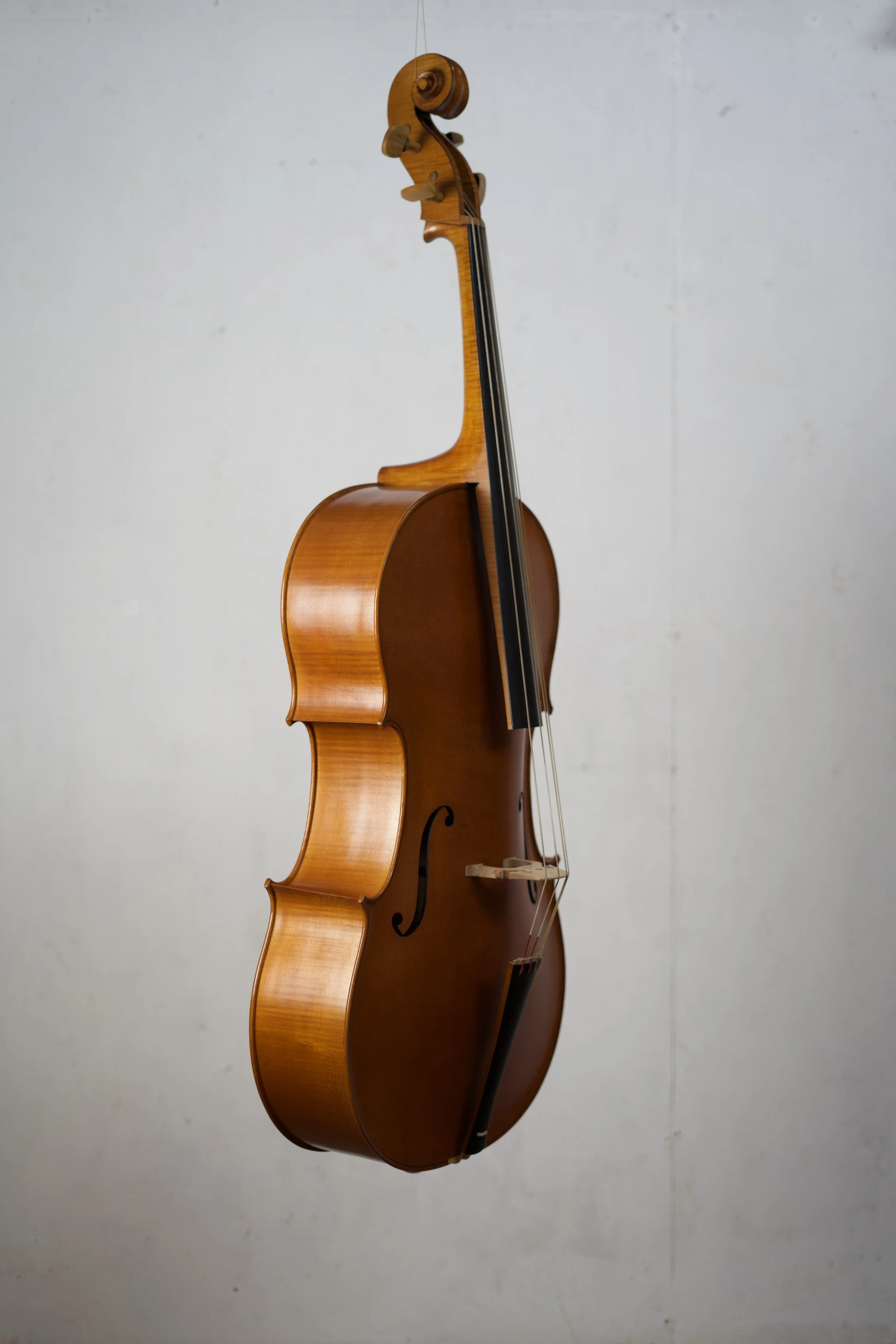A wooden cello with a light brown finish standing upright against a plain, light-colored wall.