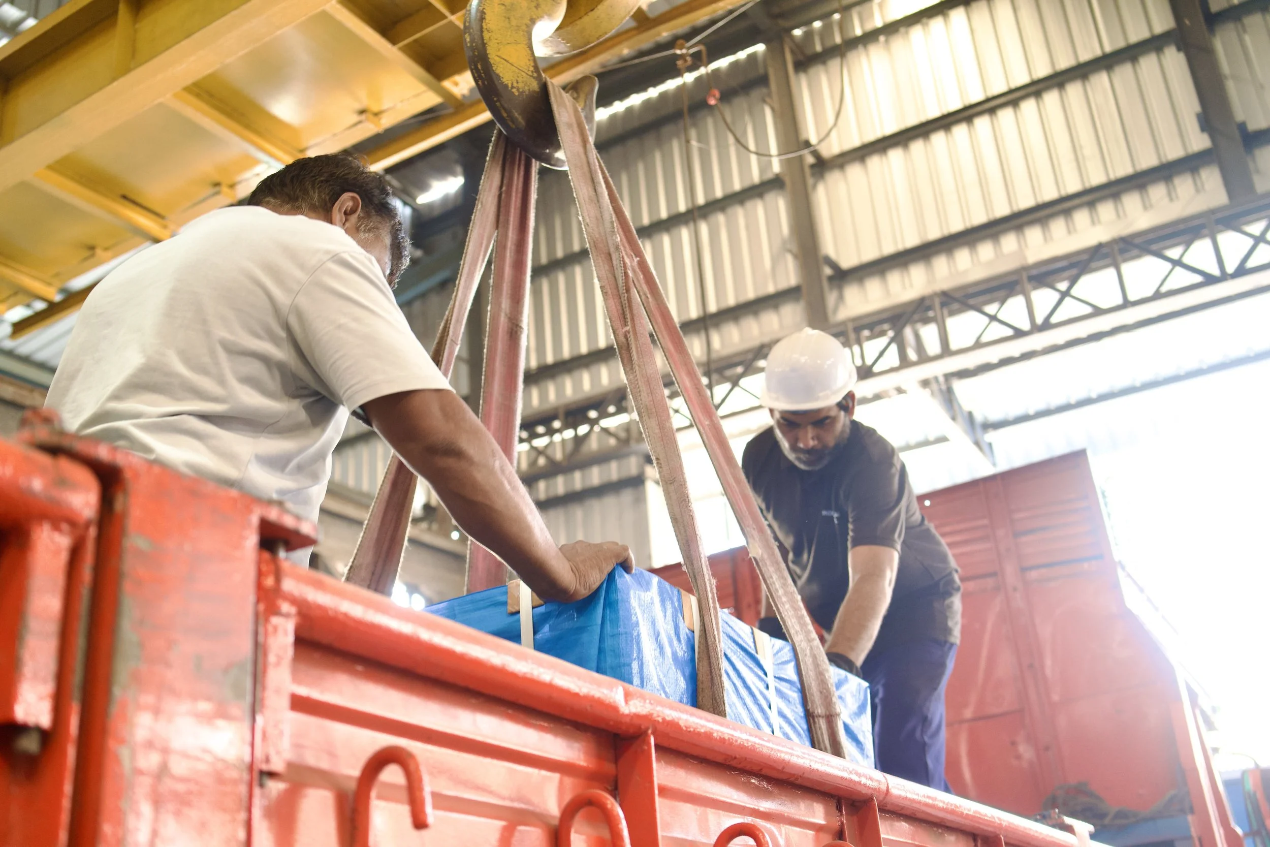 Two workers wearing hard hats and work shirts are adjusting a blue object inside a red container in an industrial setting.