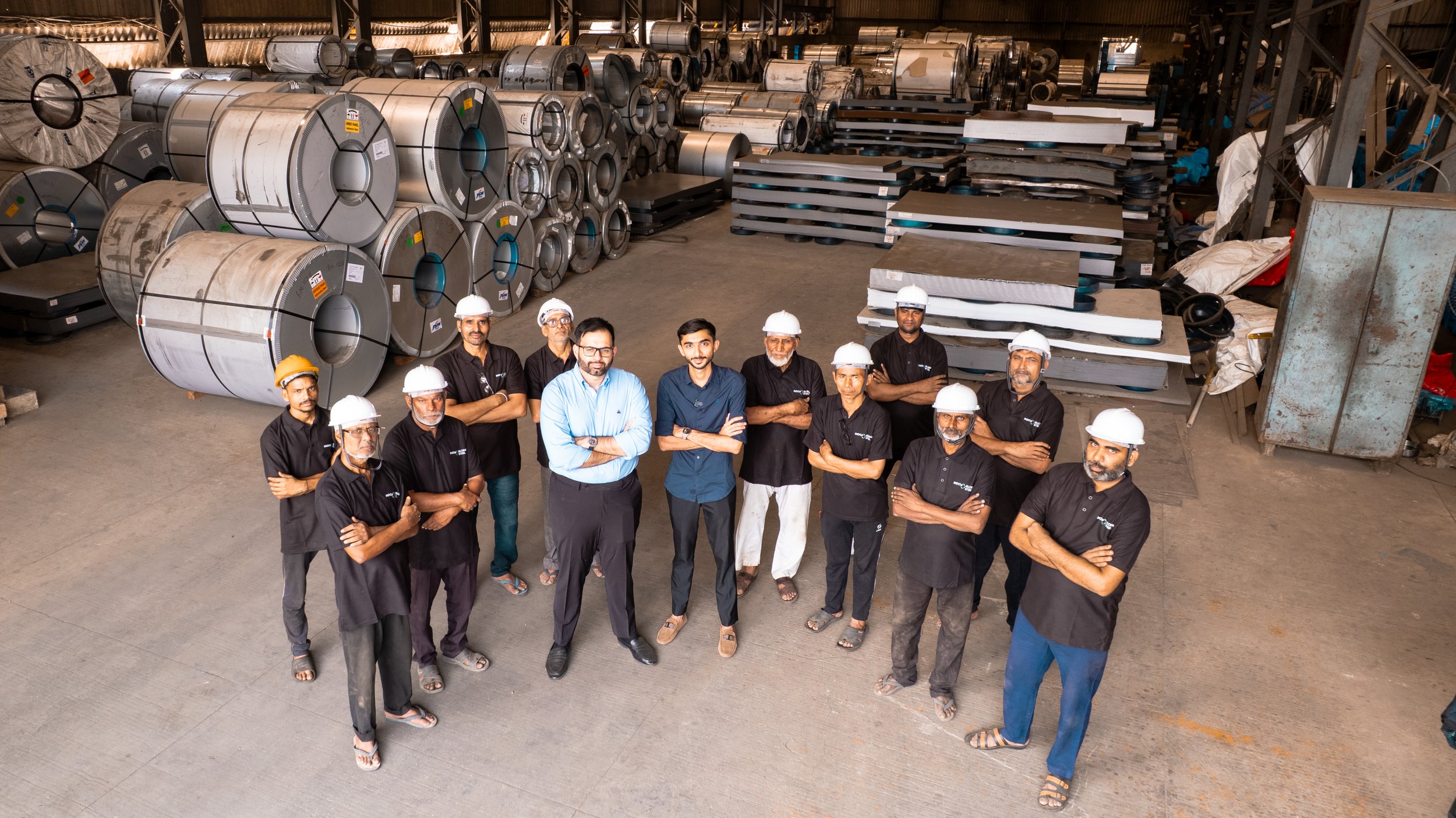Group of workers and engineers in protective helmets standing with arms crossed in a warehouse with large coils of metal and metal sheets on pallets in the background.