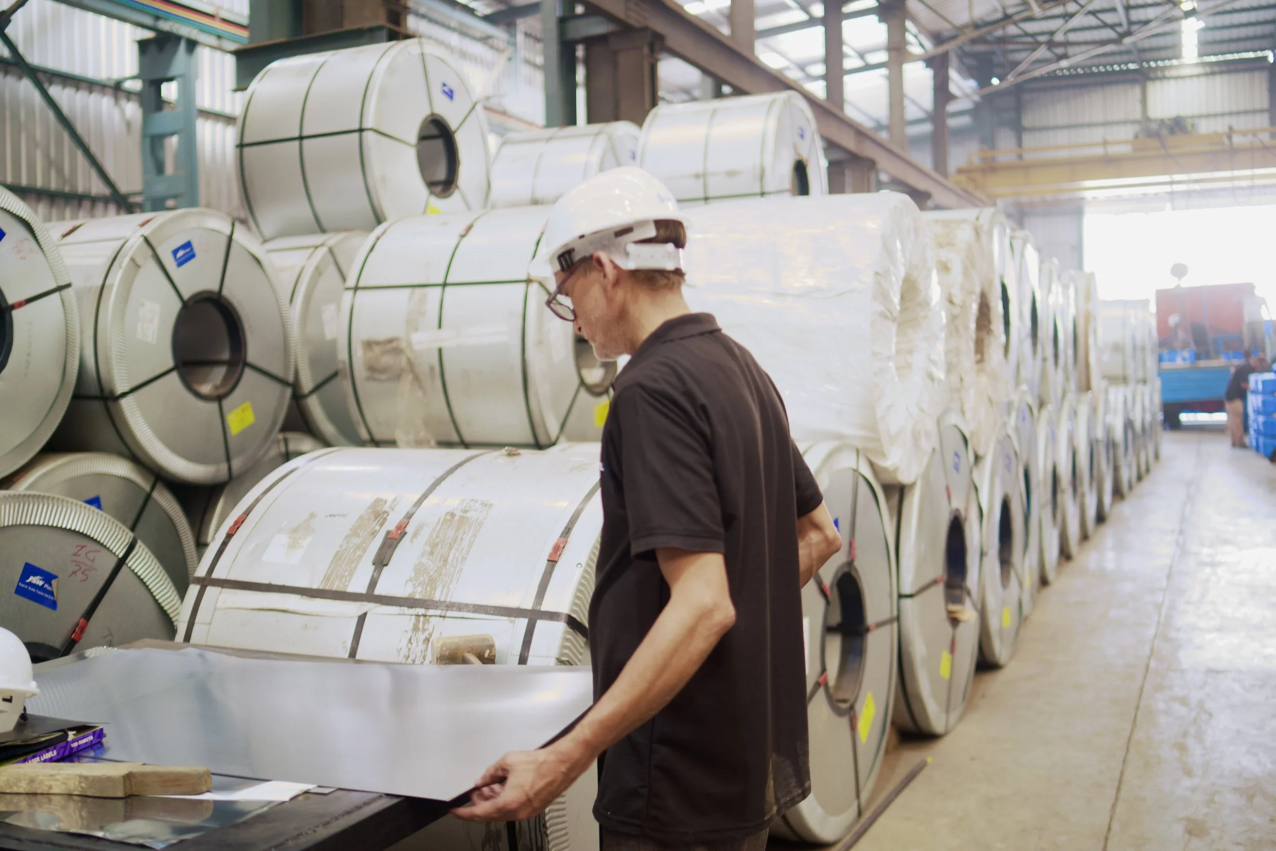 A worker in a factory wearing a safety helmet and glasses, handling a large metal duct in a warehouse with multiple rolls of insulation ductwork stacked in the background.