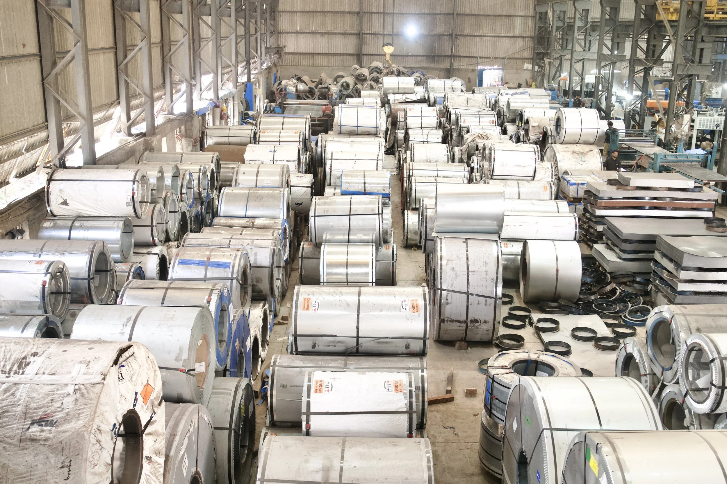 Warehouse filled with large rolls of sheet metal stacked on metal racks, with some machinery and workers visible in the background.