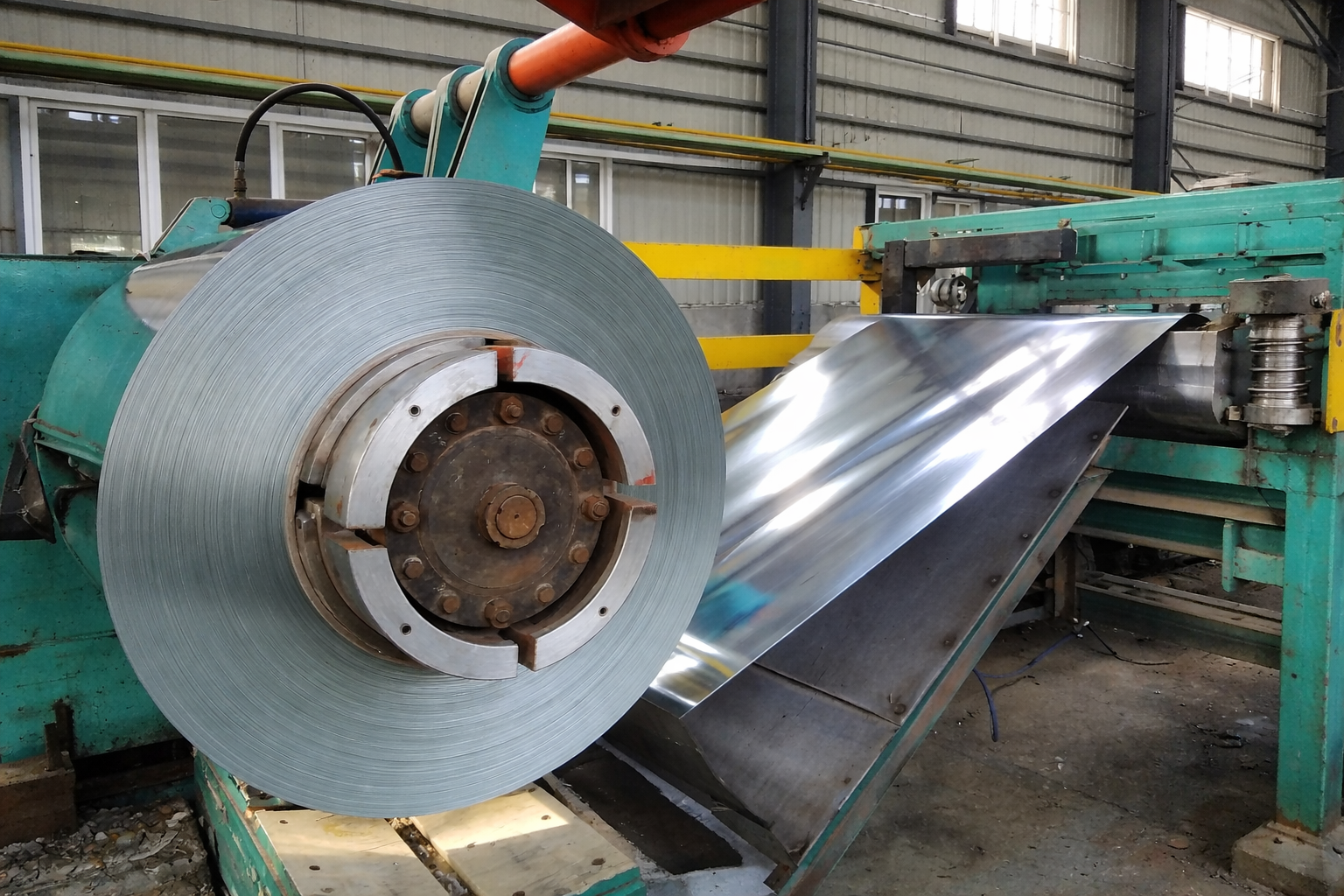 Industrial manufacturing process with a large roll of metal sheet unwinding and moving through machinery in a factory setting.