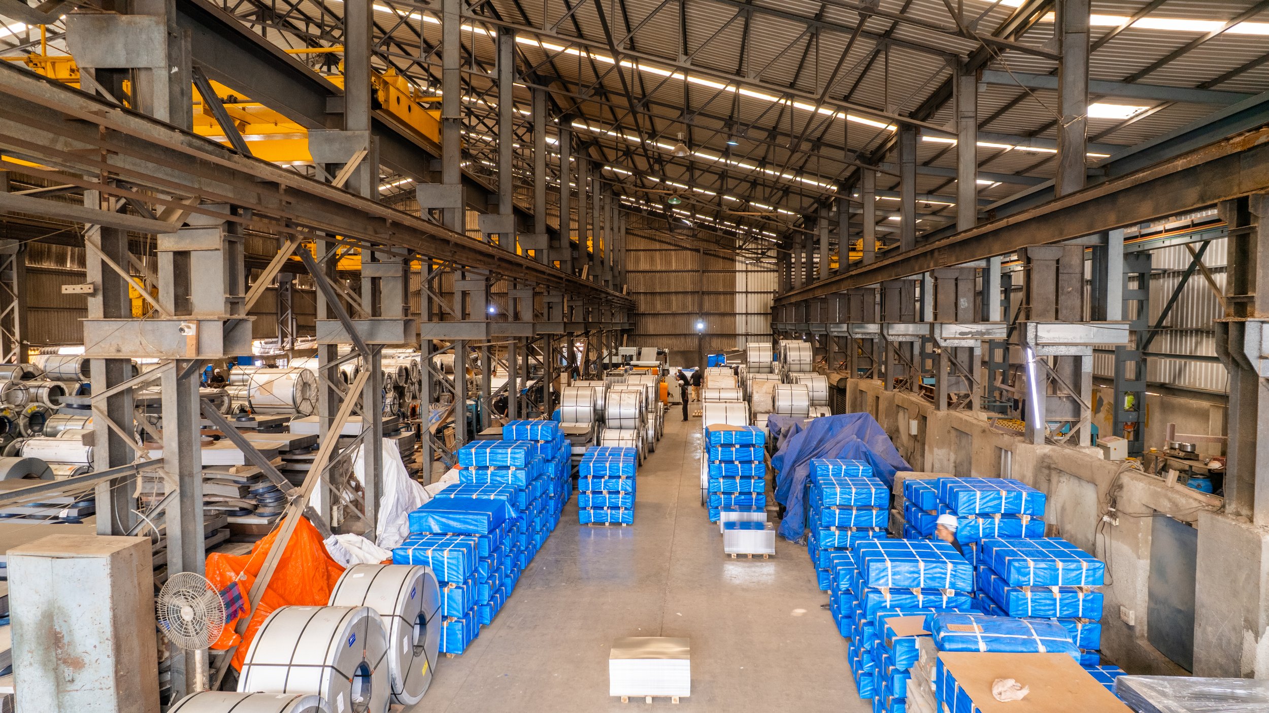 Inside a large industrial warehouse with metal beams, forklift tracks, and stacks of metal coils and blue wrapped packages on pallets.
