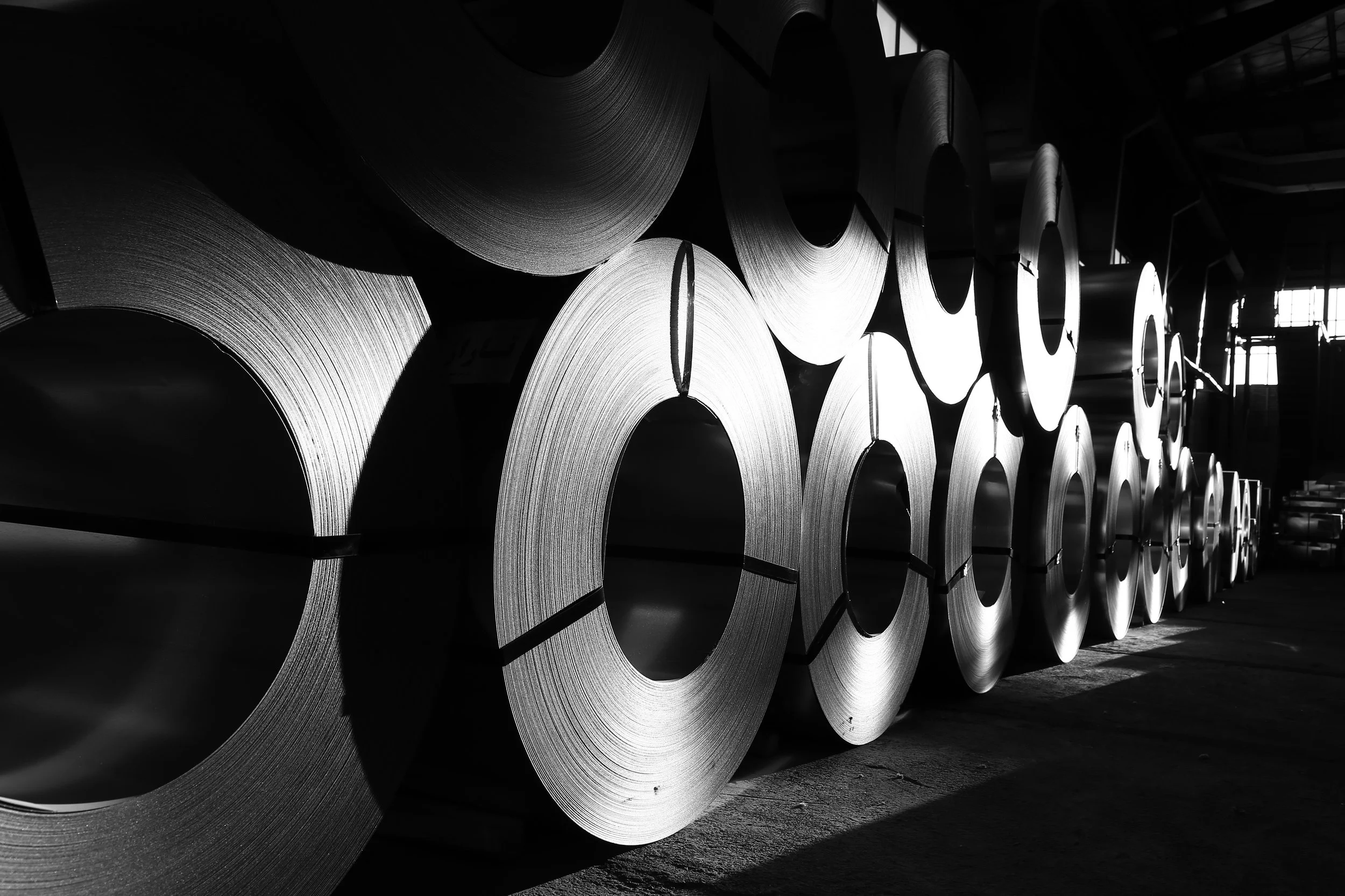 Stacks of large steel coils stored in an industrial warehouse, with light and shadow creating a high-contrast black-and-white scene.