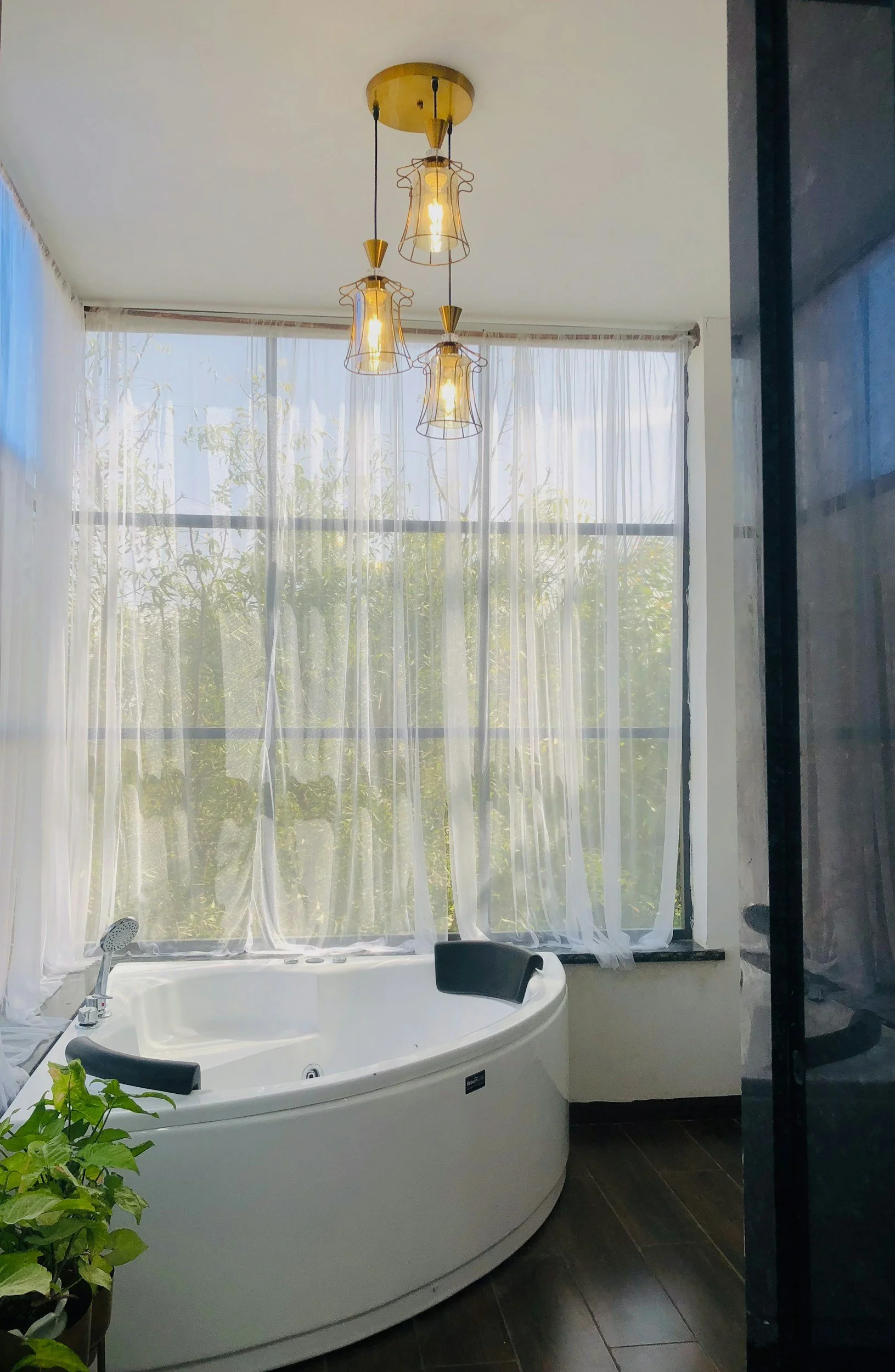 A bathroom with a large bathtub near a window with sheer white curtains and a hanging light fixture.