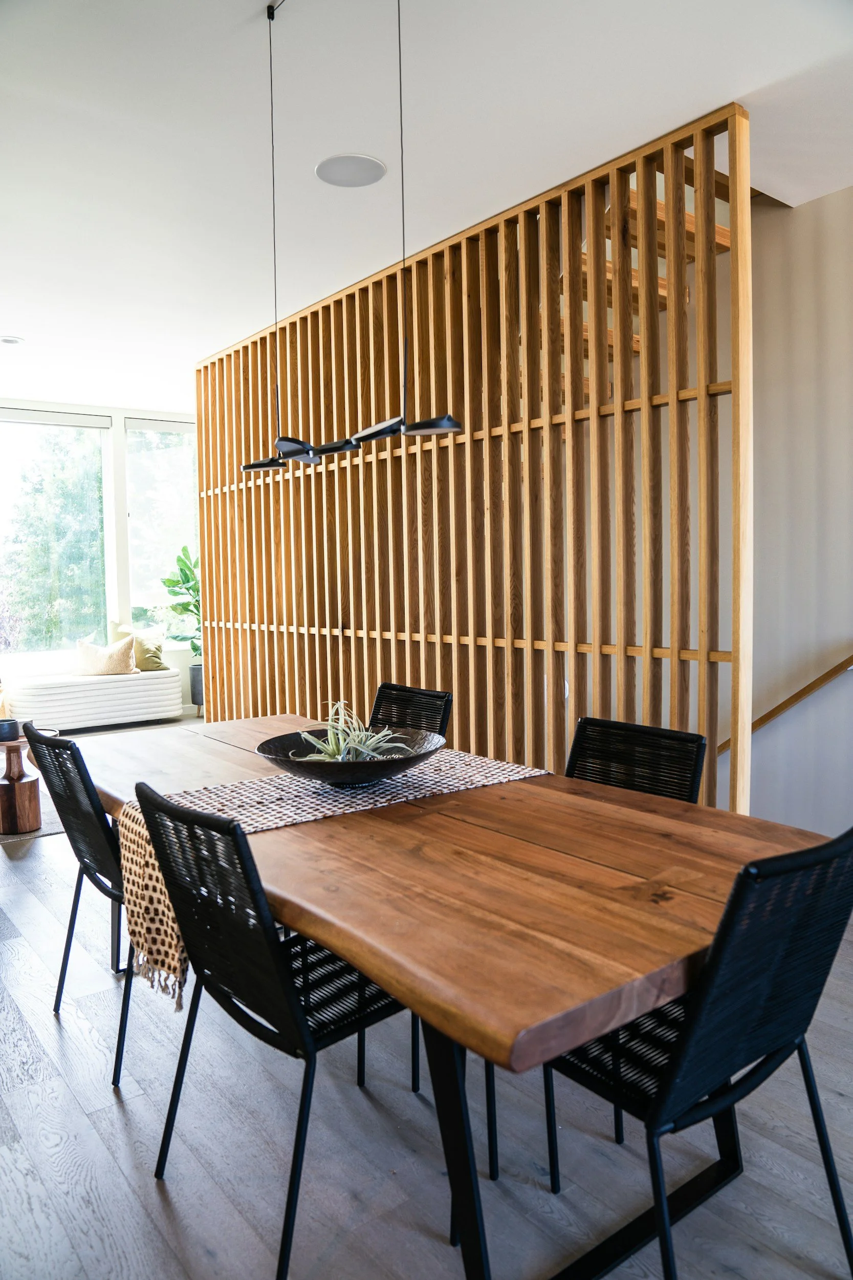 A dining room with a wooden table and black chairs, a wooden partition, and a window with a sofa and plants outside.