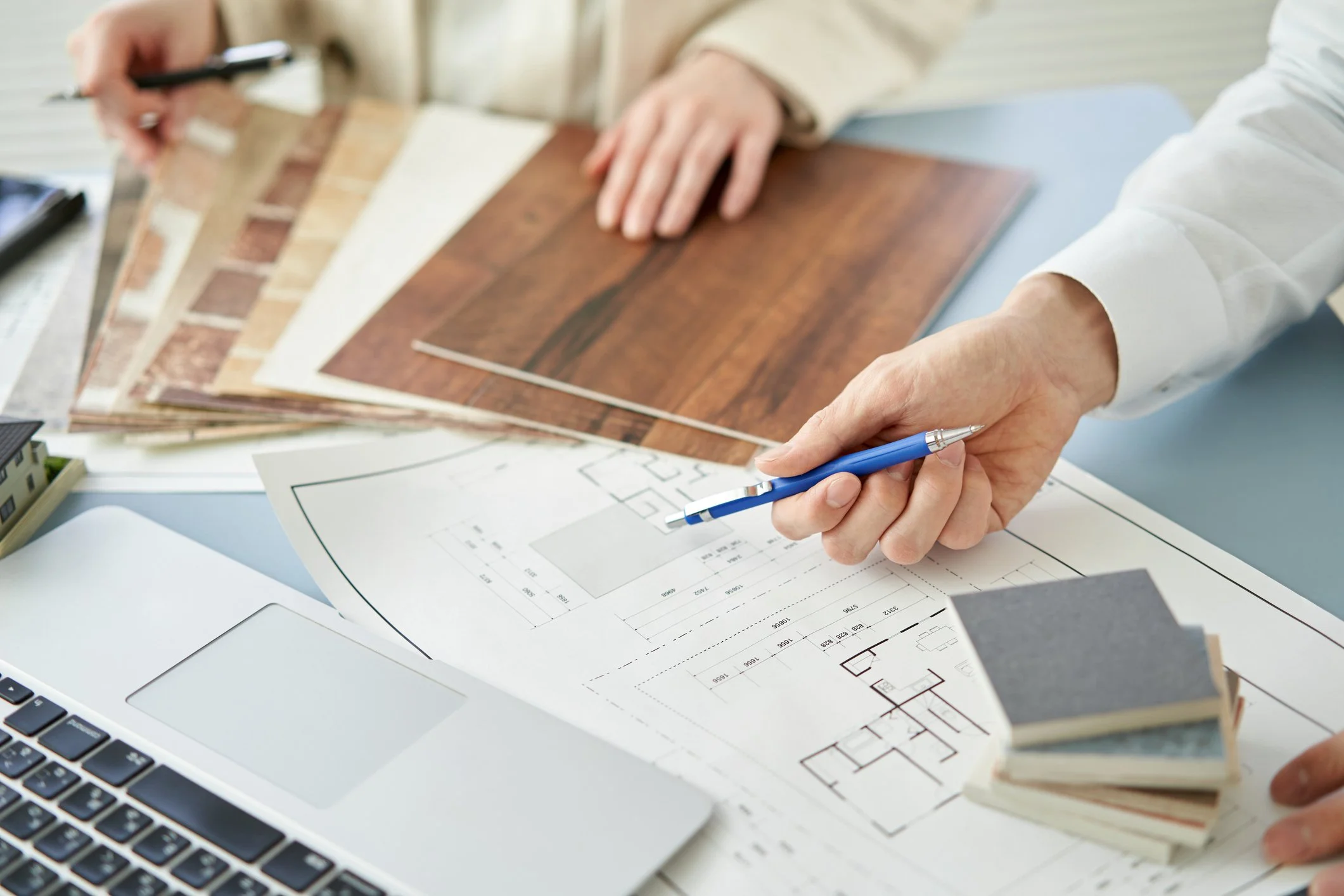 People examining a sample of wood flooring with design plans and a laptop on the table.