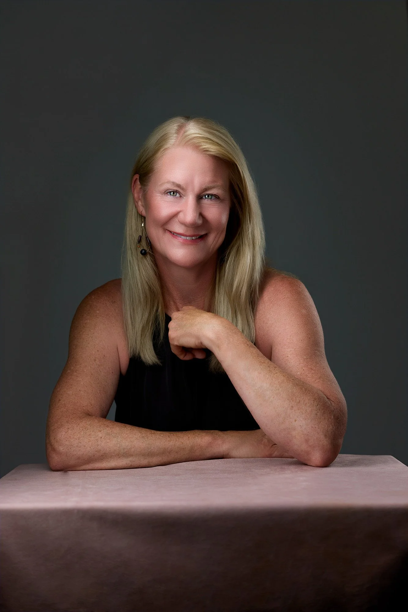 Portrait of Teresa Petersen, a smiling blonde woman with blue eyes, wearing a black sleeveless top and dangling earrings, sitting at a light-colored table against a dark gray background.