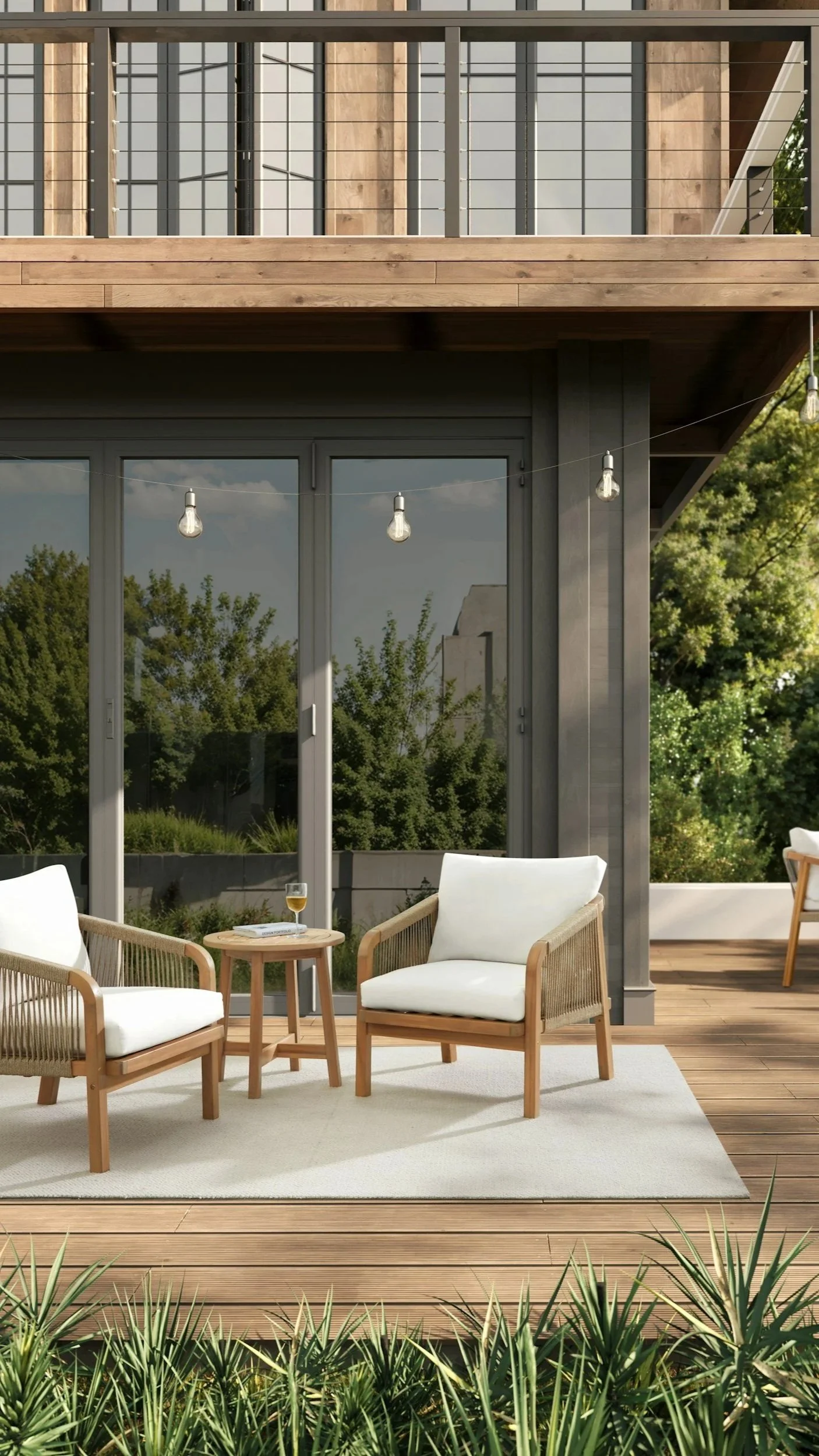 Outdoor patio with two white cushioned armchairs and a small round wooden table with a glass of wine, in front of large glass sliding doors and greenery in the background.