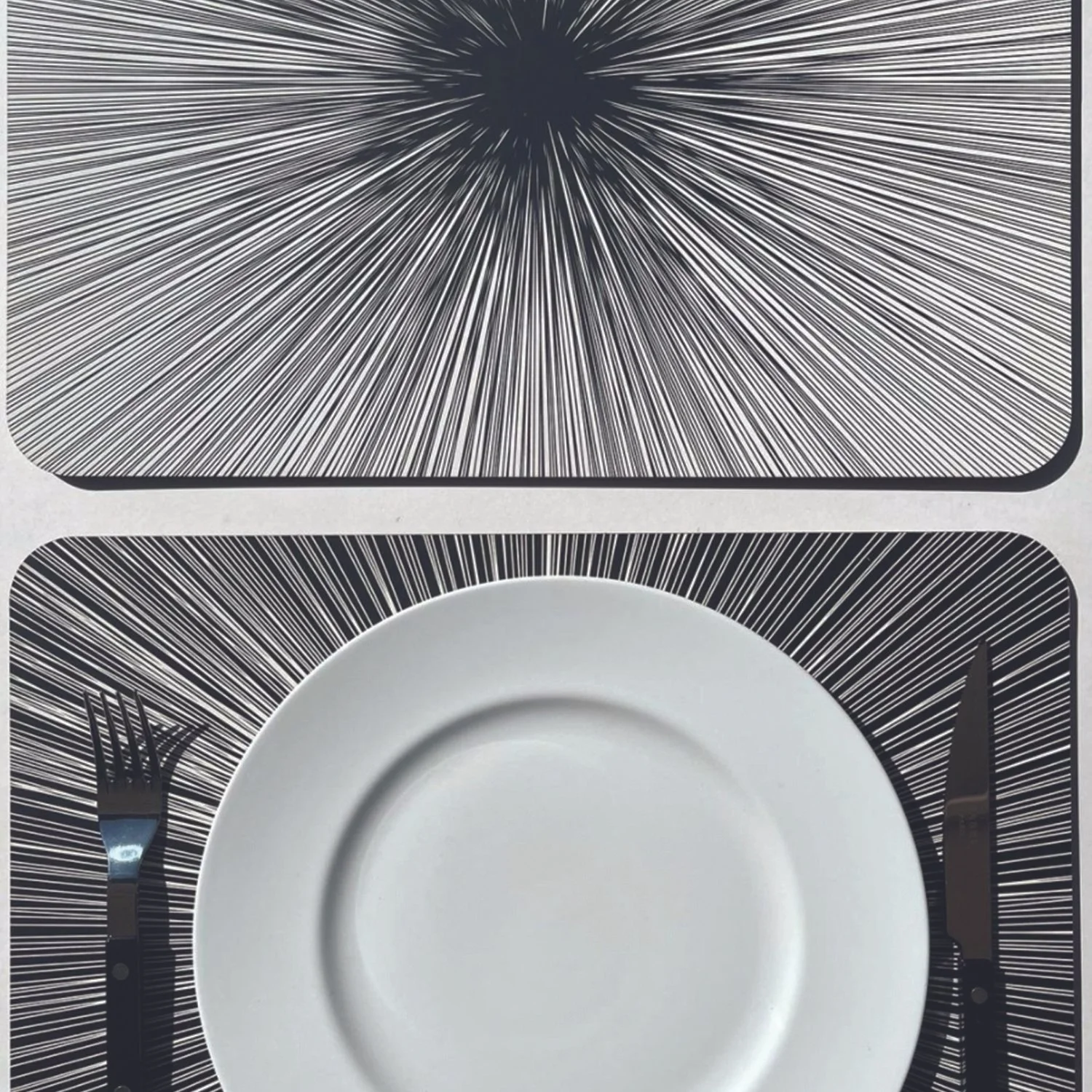 A table setting with black and white radiating line placemats underneath a white plate, surrounded by a fork on the left and a knife on the right.