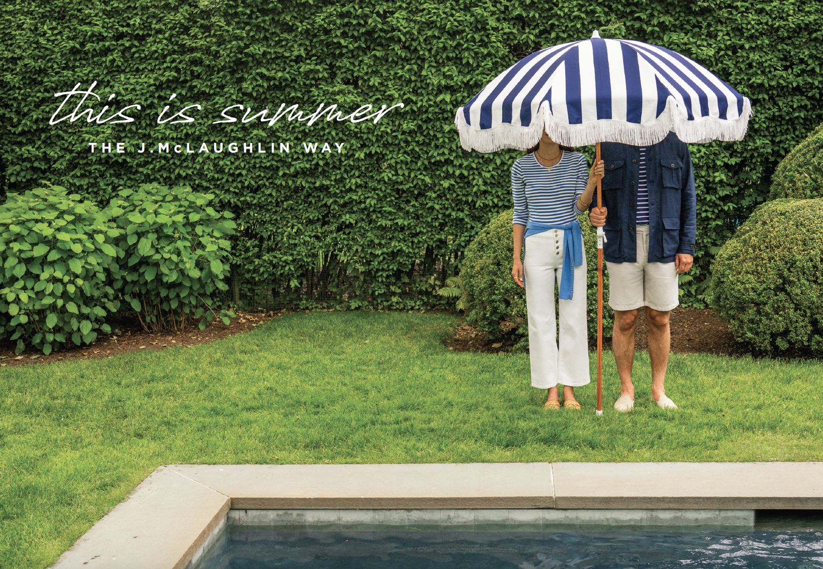 A woman and a man standing on a lawn, holding a large blue and white striped umbrella. The woman is wearing a striped shirt and white pants, and the man is wearing a blue jacket and shorts. There are bushes and a hedge in the background, and the scen