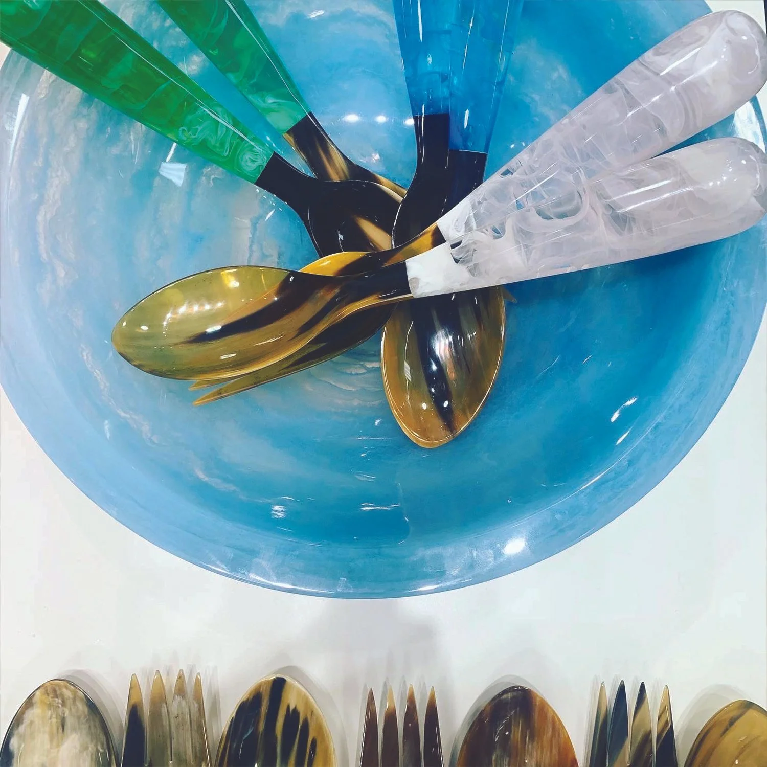 Colorful spoons inside a large blue glass bowl, with additional utensils at the bottom of the image.