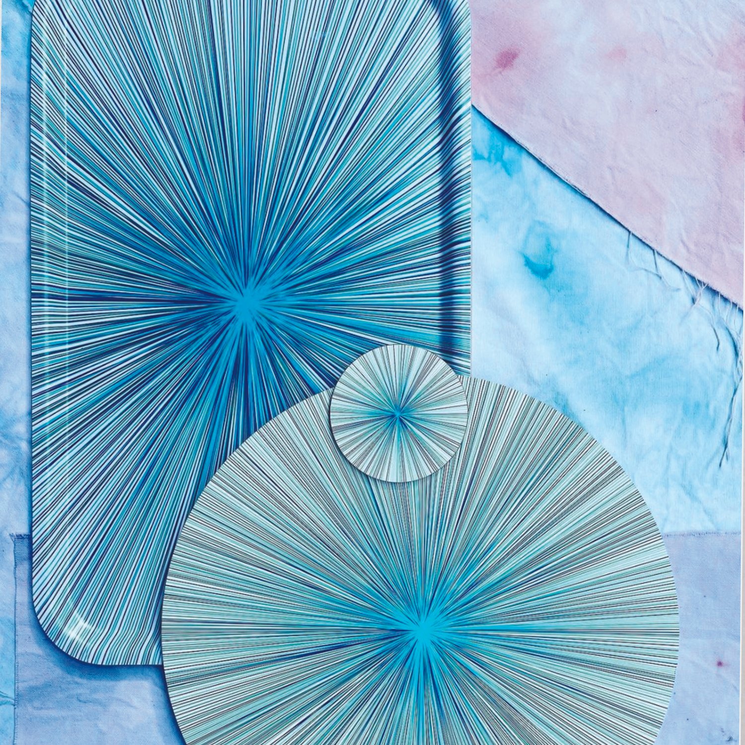 Three paper circles with blue and black radial line patterns on a fabric surface, with a large sheet of paper with a similar pattern in the background.