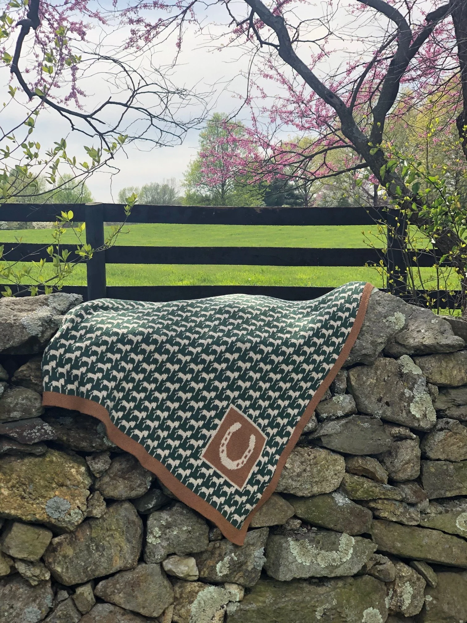 A patterned blanket with a horseshoe symbol draped over a stone wall in a rural setting with green grass, trees, and pink blossoms in the background.